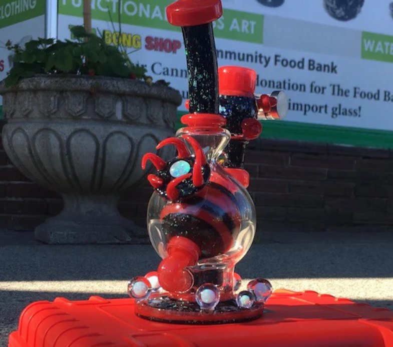 A decorative glass bong with red and black accents sitting on a red surface outdoors, with a large plant pot and a parking lot barrier in the background.