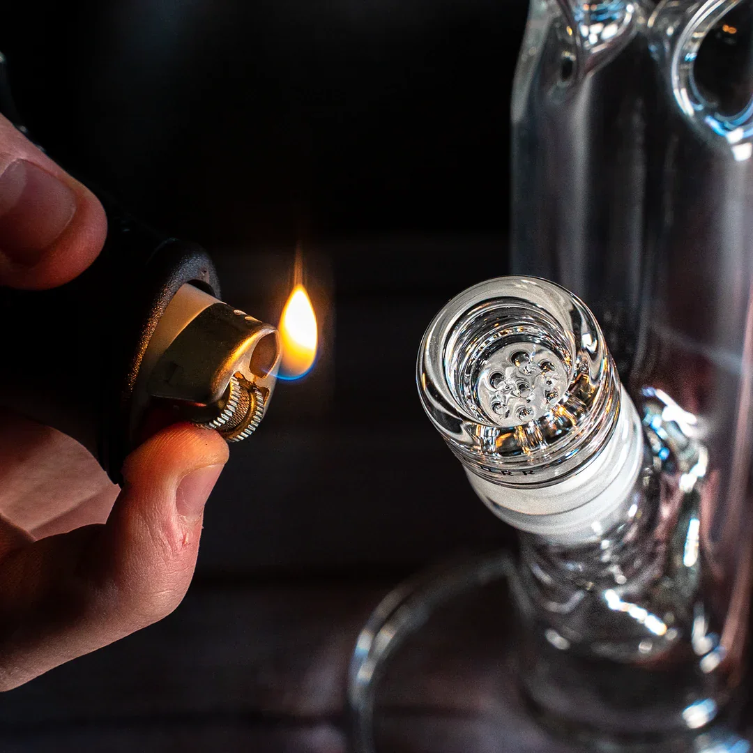 A hand holding a lighter with a flame, igniting the top of a glass water pipe or bong.