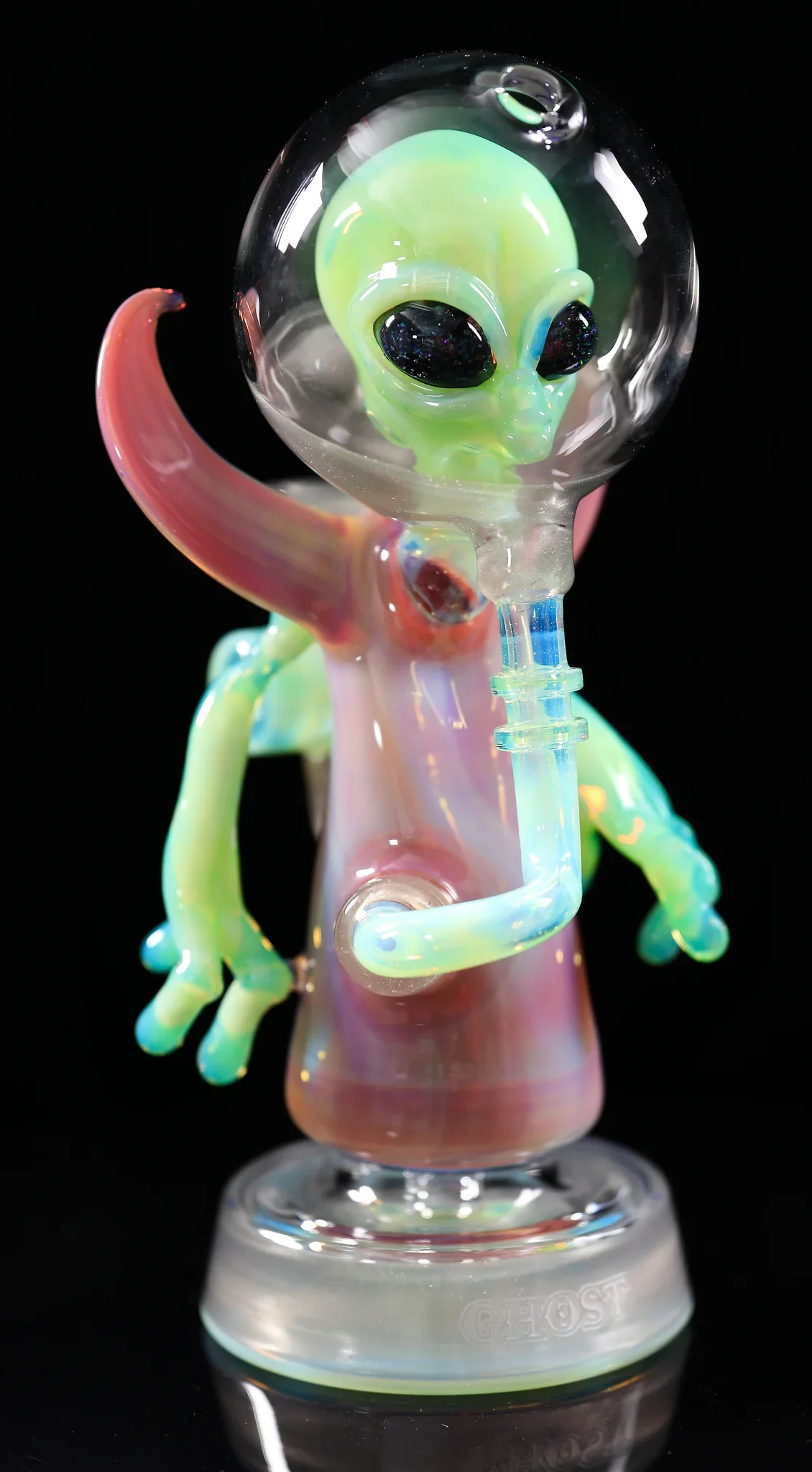 Colorful glass alien figure with a large head, black eyes, and a small body, holding a glass pipe, against a black background.