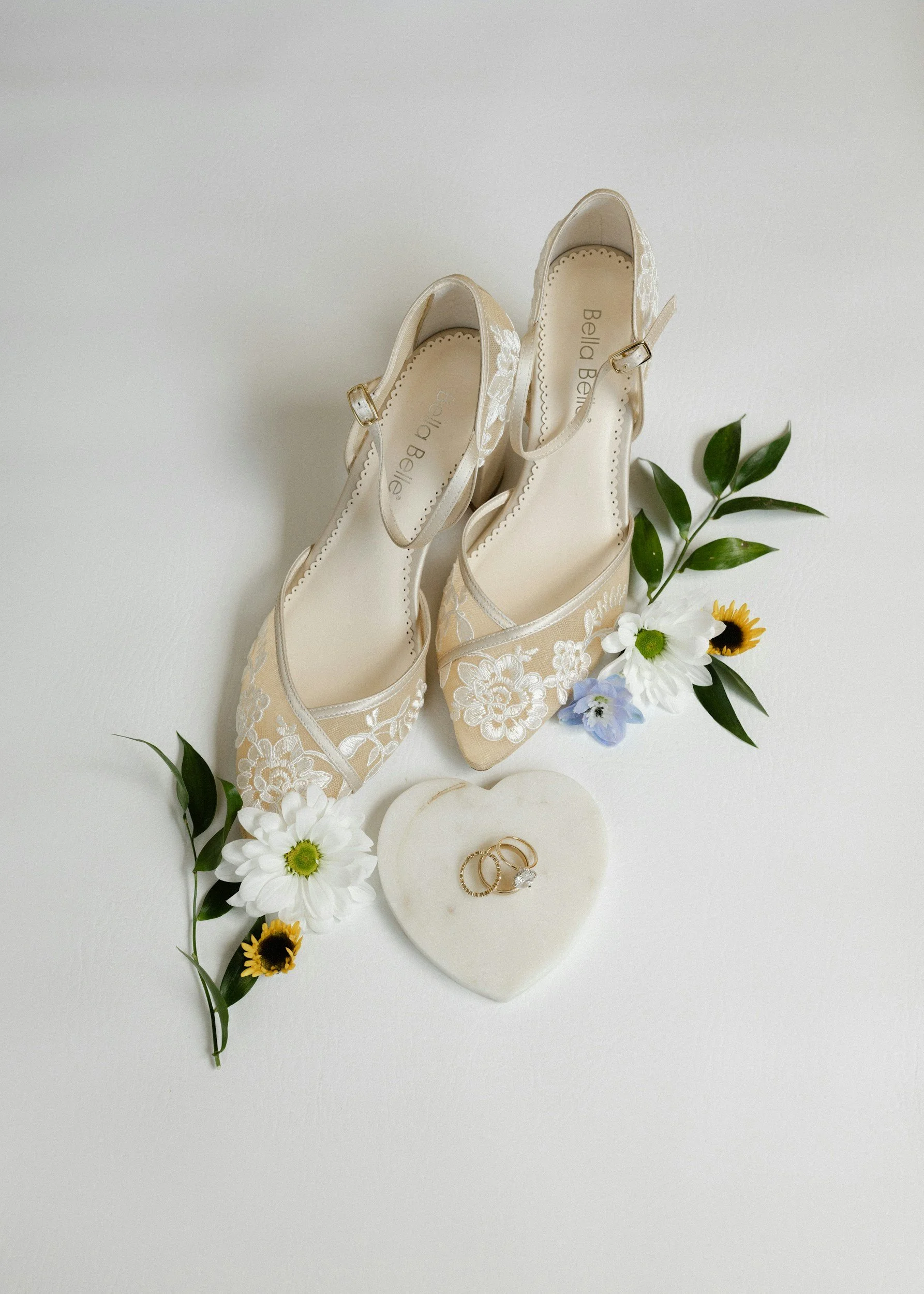 Ein Paar weiße Brautschuhe mit Spitzenbordüre, zwei Eheringe auf einer Herz-Dekoration, umgeben von weißen, gelben und lilafarbenen Blumen, auf einem weißen Hintergrund.