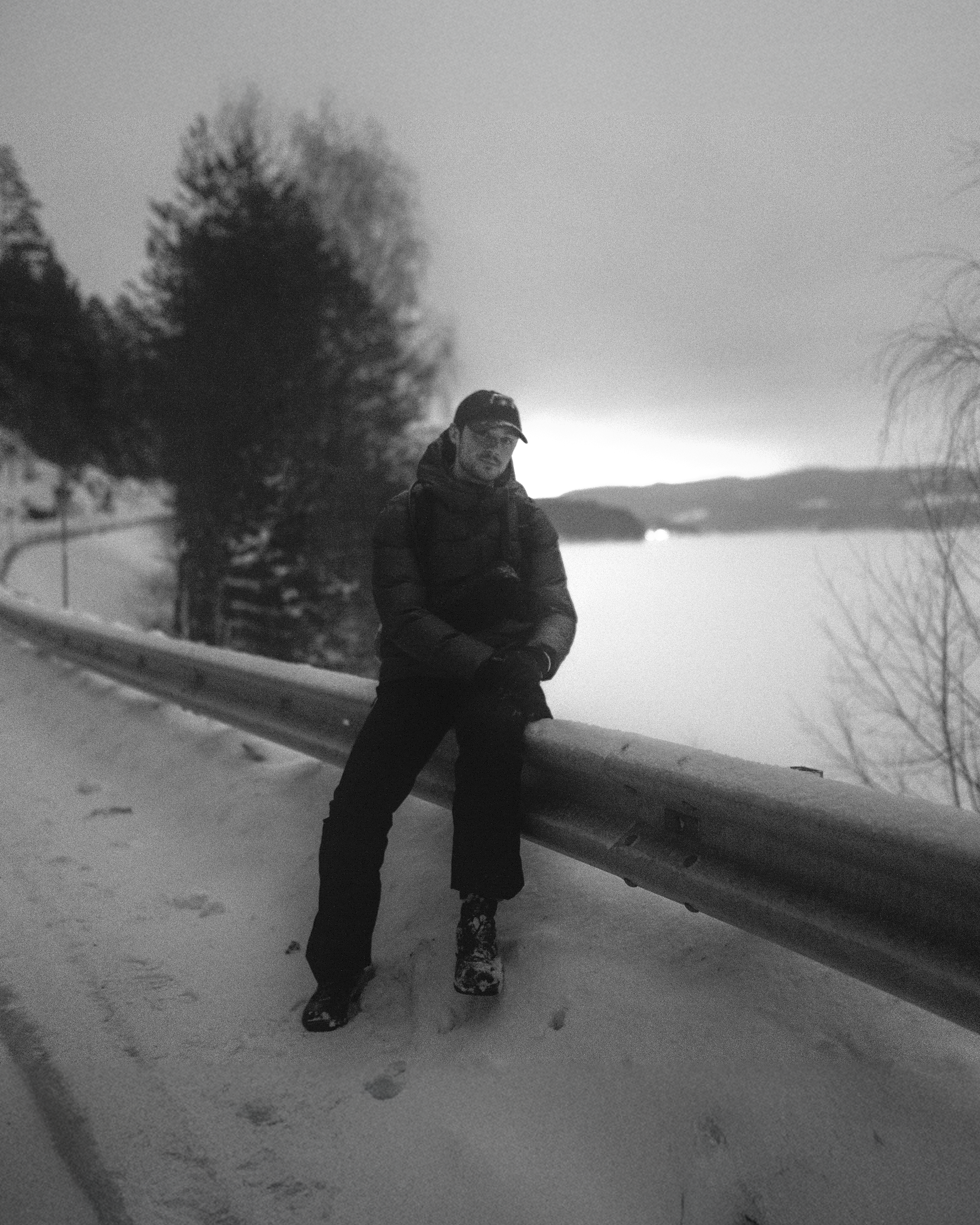 A person wearing winter clothing leaning on a guardrail along a snowy road with trees and a large body of water in the background, in black and white.