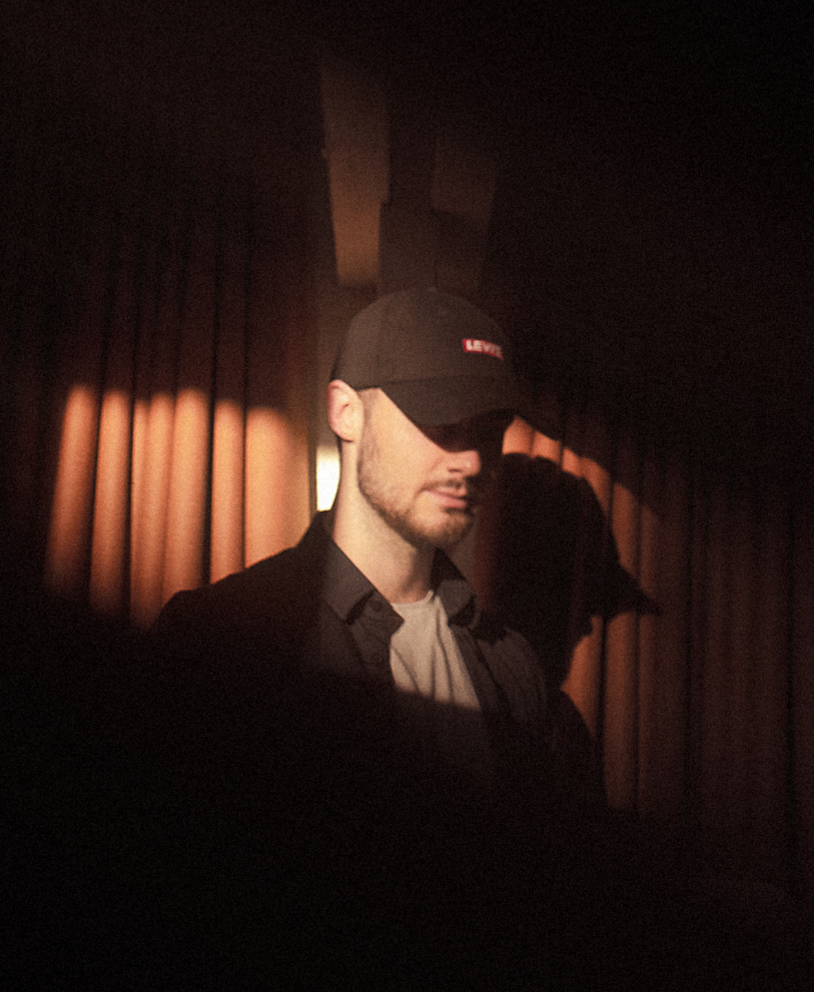A man wearing a black cap with a red logo, a black jacket, and a light-colored shirt, standing against a backdrop of brown curtains with warm lighting.