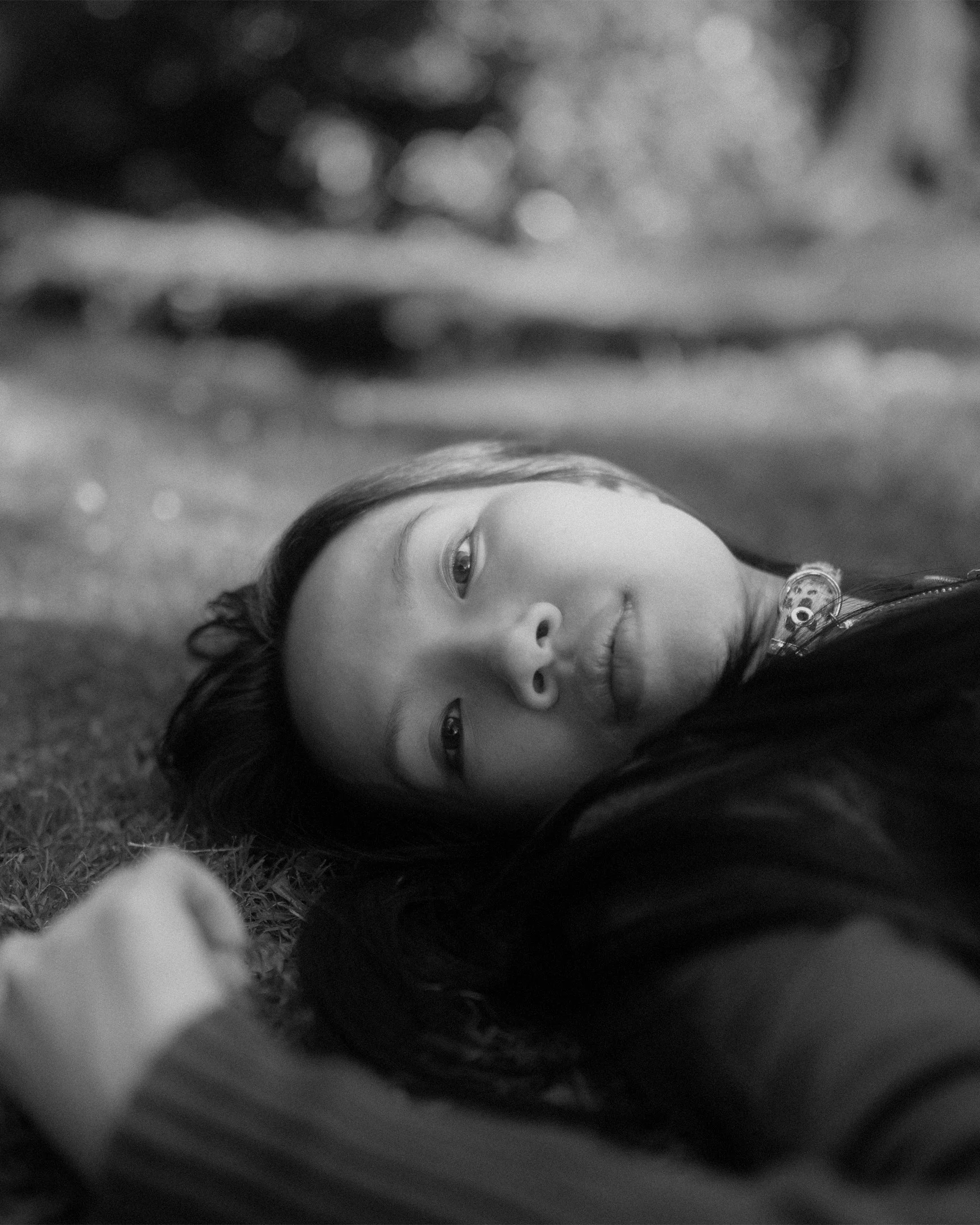 A young girl lying on her side on the grass, with long dark hair, looking up with a contemplative expression. The background is blurry with bokeh effects.