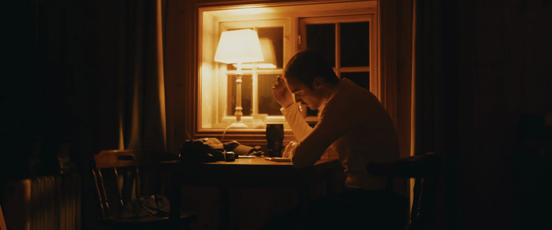 A young man sitting at a table in a dimly lit room, resting his head on his hand with a contemplative expression, illuminated by a warm table lamp.