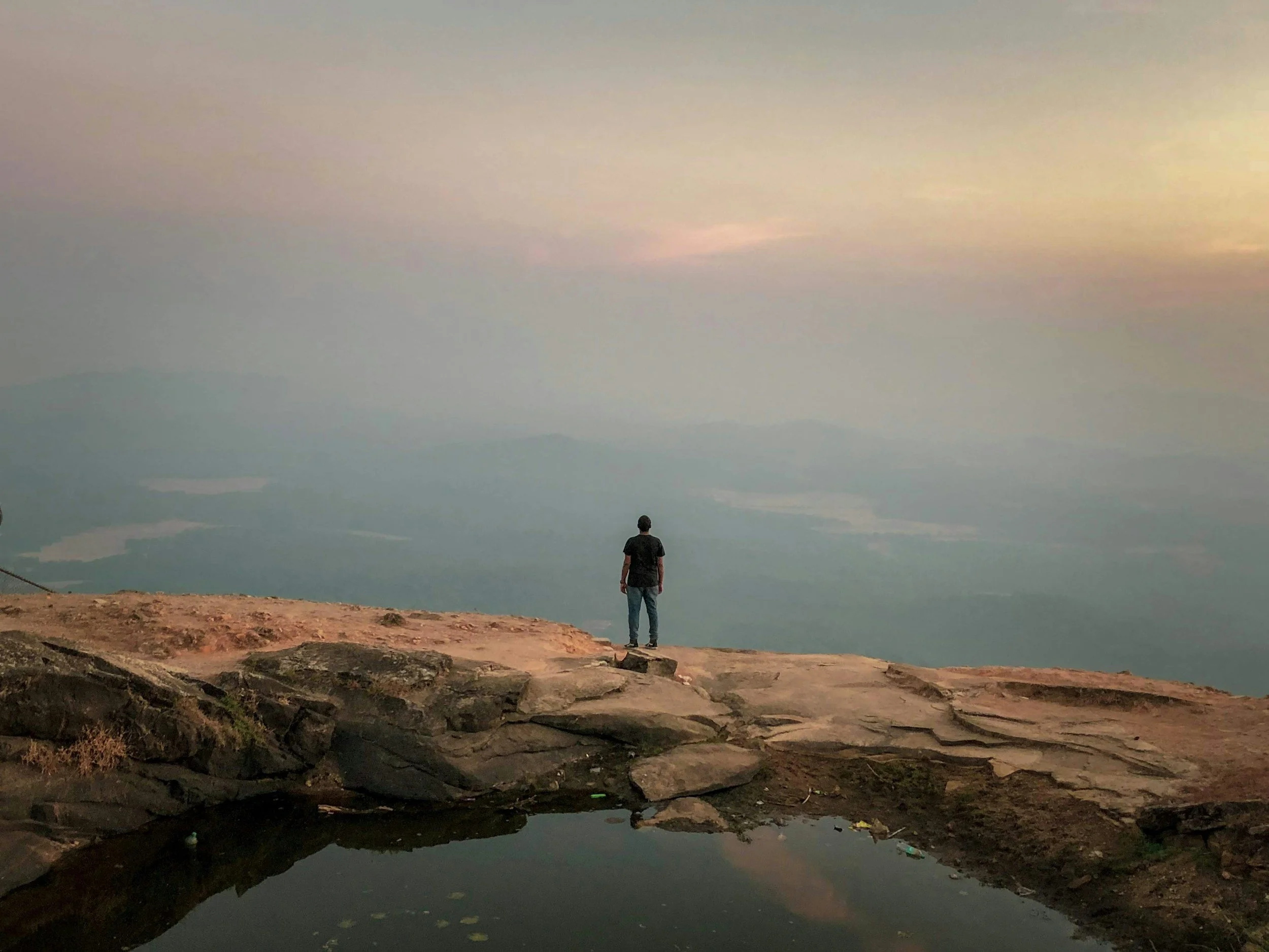 A person standing on a rocky cliff overlooking a vast landscape with water bodies and mountains at sunset.