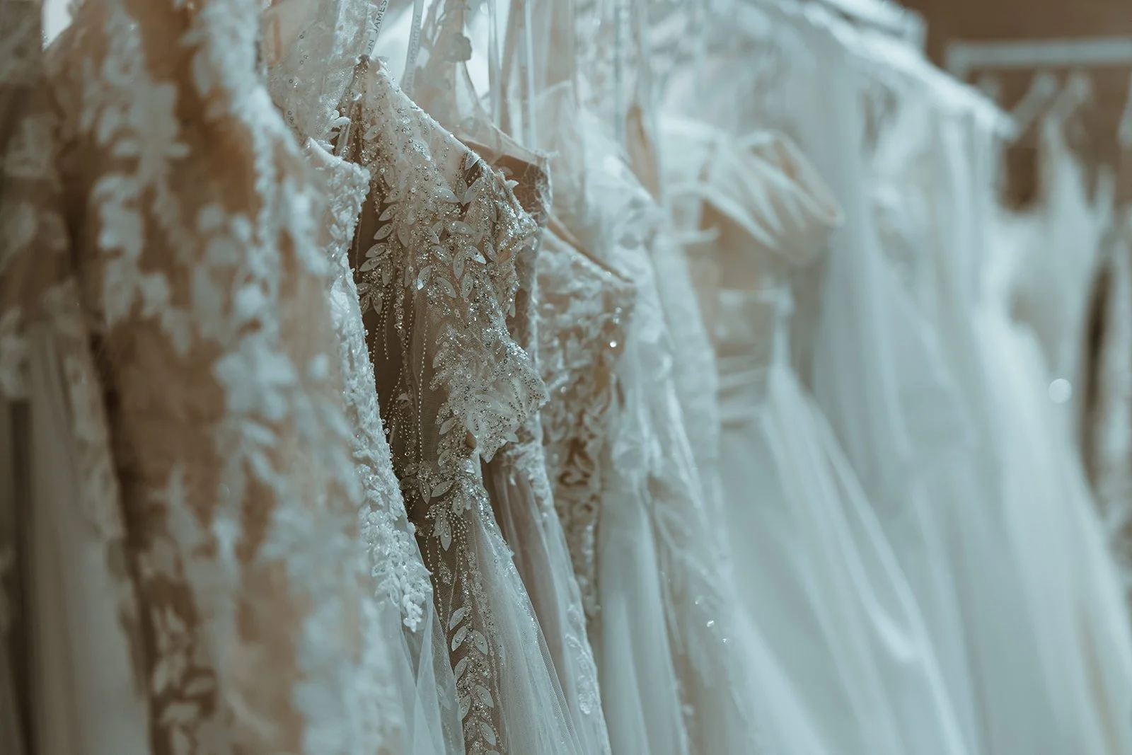 A row of wedding dresses hanging on a rack, featuring intricate lace and beadwork designs.