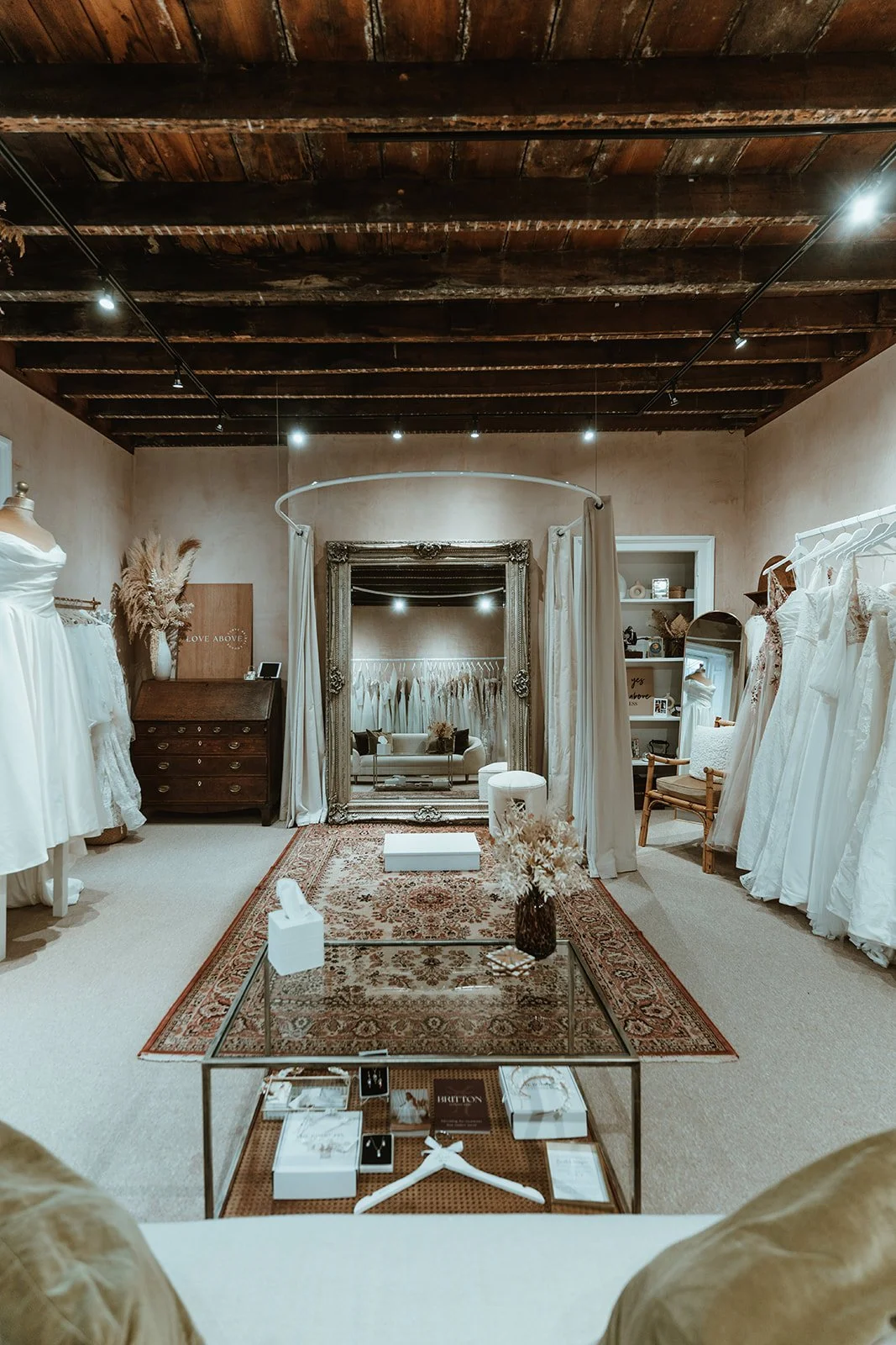 Interior of a boutique bridal shop with white wedding dresses on racks, a large ornate mirror, and a cozy seating area reflected in the mirror.