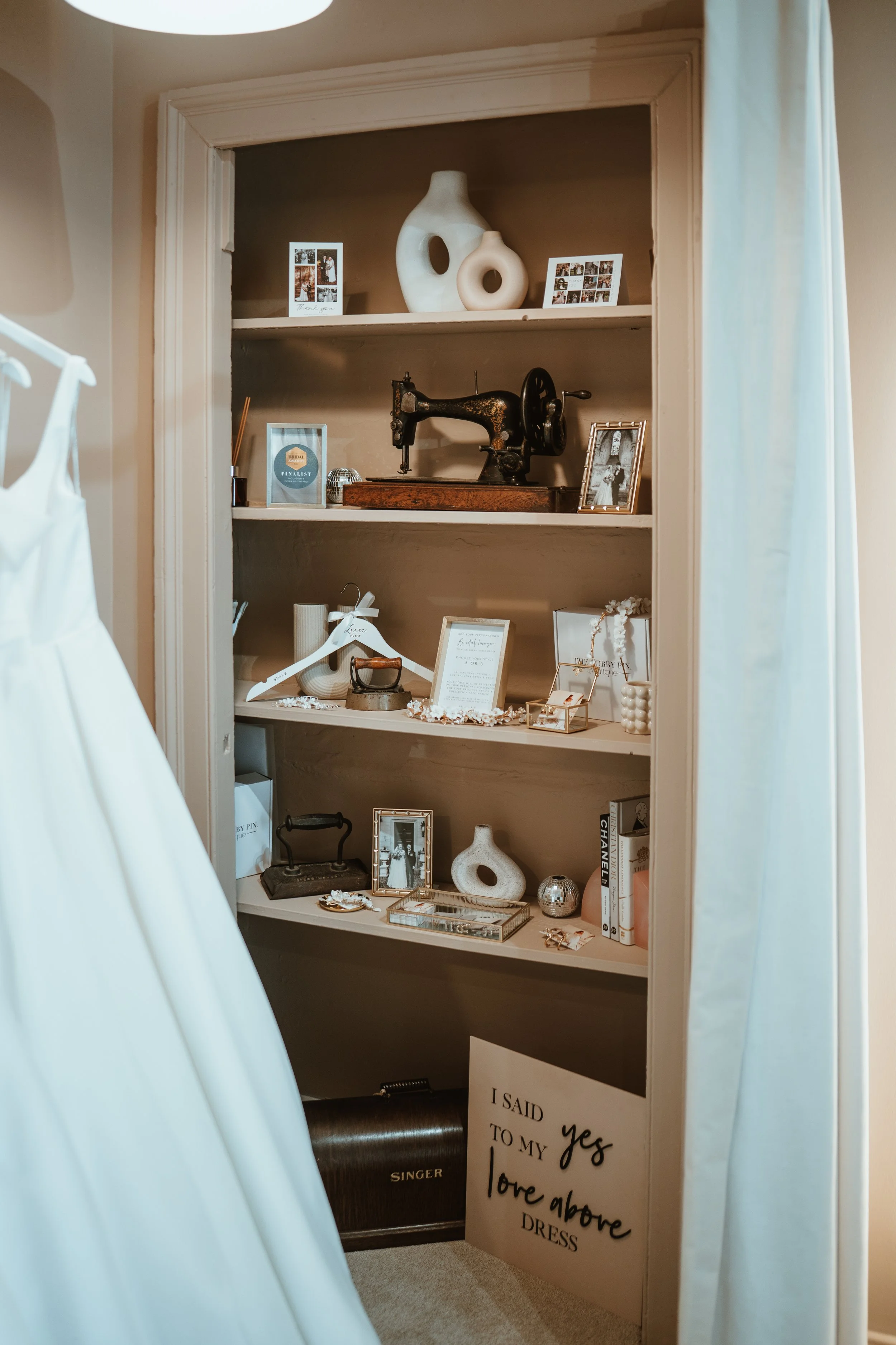 A built-in bookshelf with decorative items including vases, framed photos, an antique sewing machine, a mirror, books, and various ornaments, partially obscured by white fabric in the foreground.