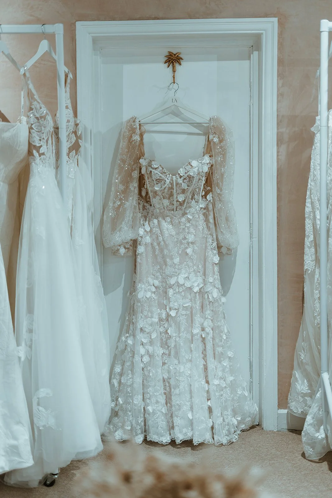 A wedding dress with floral lace and tulle details hanging on a white hanger on a door frame, surrounded by other white wedding dresses on racks.