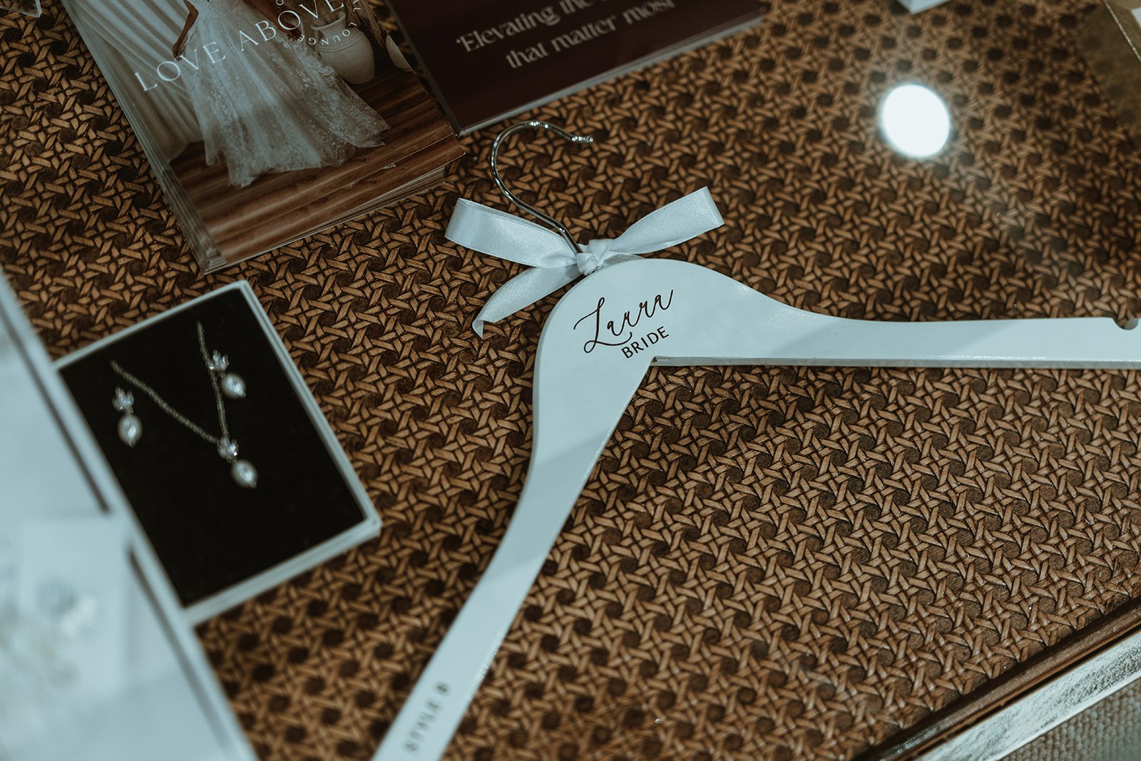 A white hanger with the name 'Laura' and the word 'Bride' written on it, placed on a patterned surface surrounded by jewelry boxes containing earrings and a necklace, with a wedding dress magazine nearby.