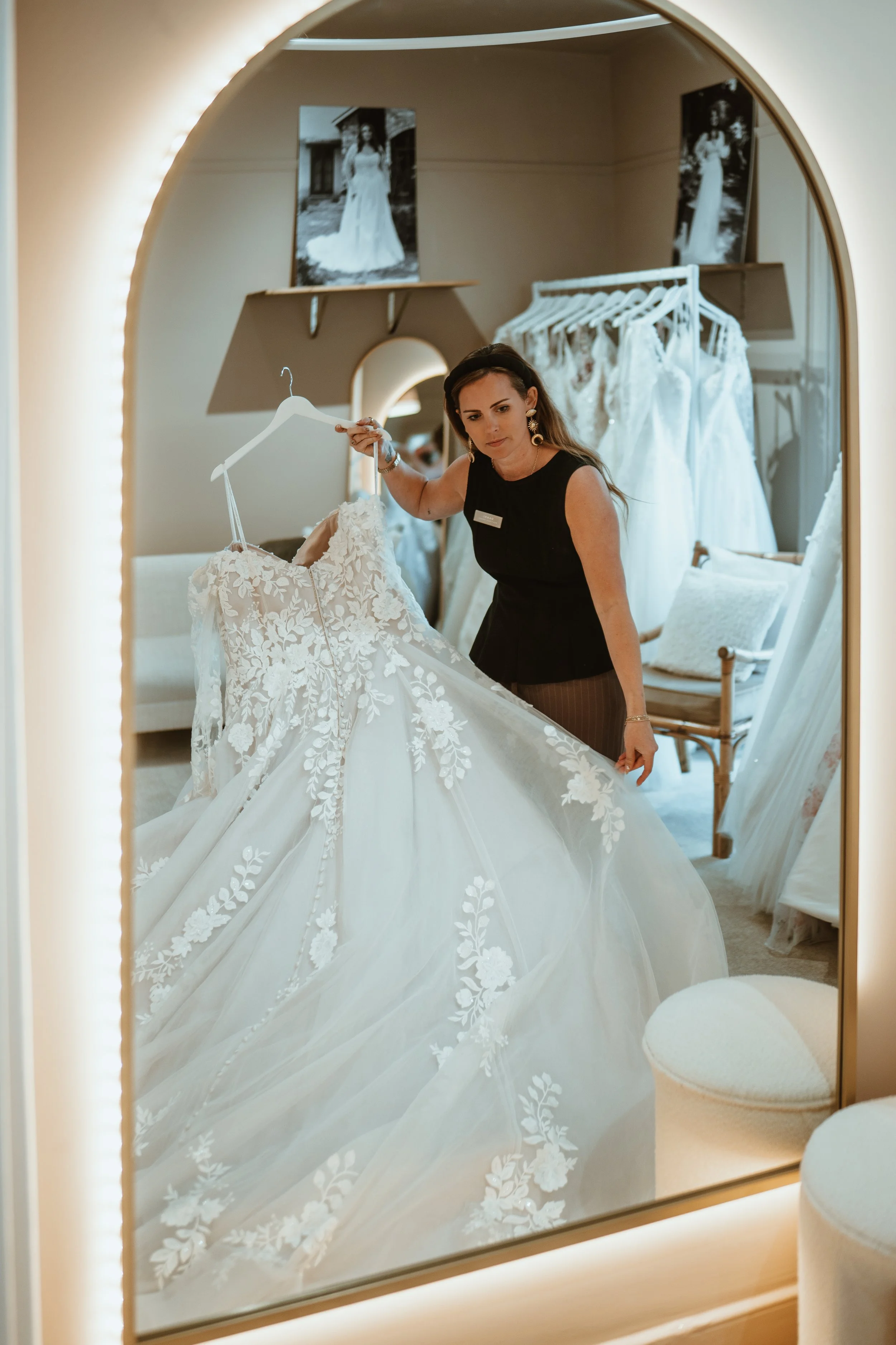 A woman trying on a wedding dress in a fitting room, looking at the dress while holding it on a hanger. The reflection shows two photographs of a bride in a wedding gown hanging on the wall behind her. There are also white wedding dresses hanging on 