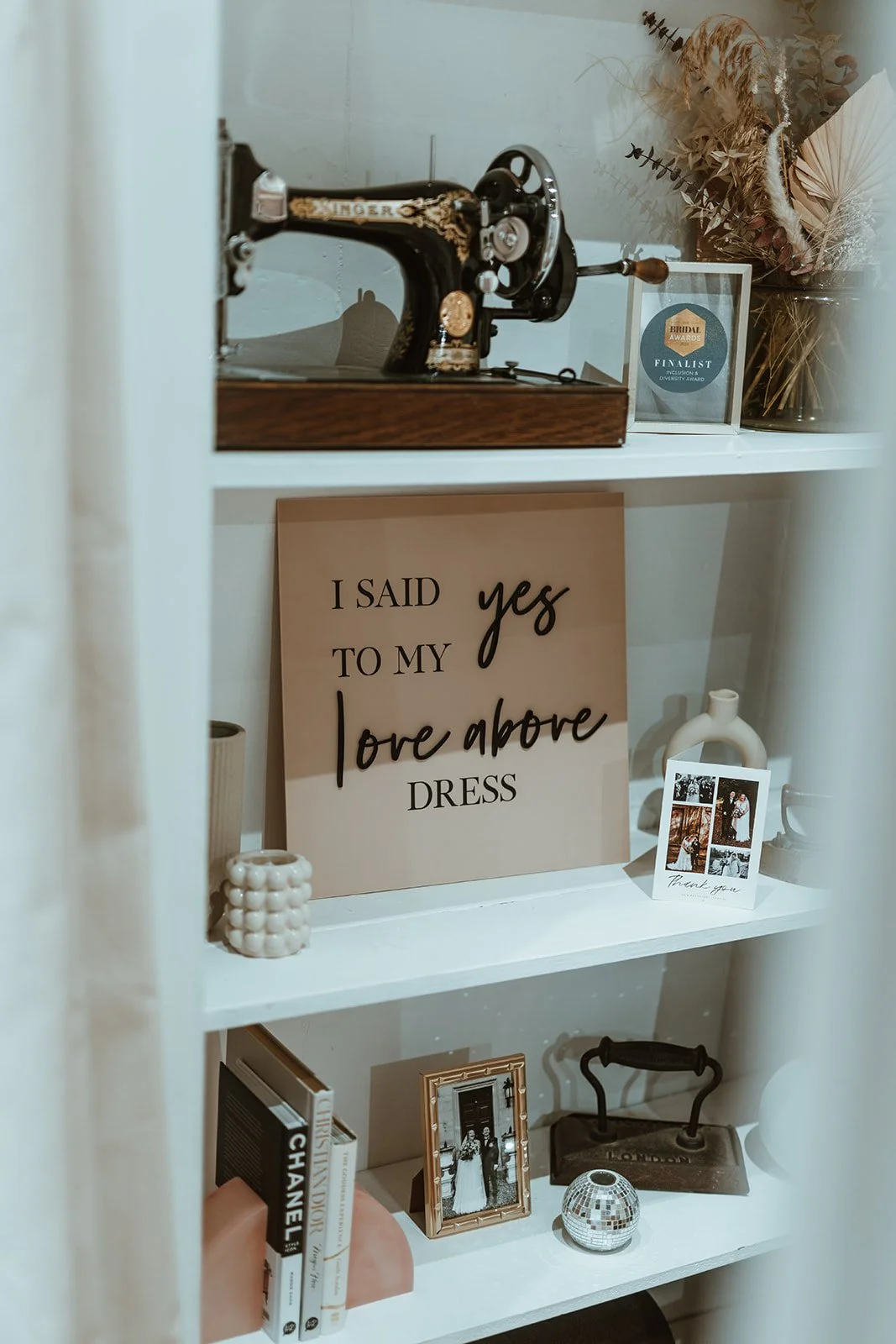 A display shelf with vintage sewing machine, dried floral arrangement, wedding photo frame, decorative sign reading 'I said yes to my love above dress', small books, and decorative items.