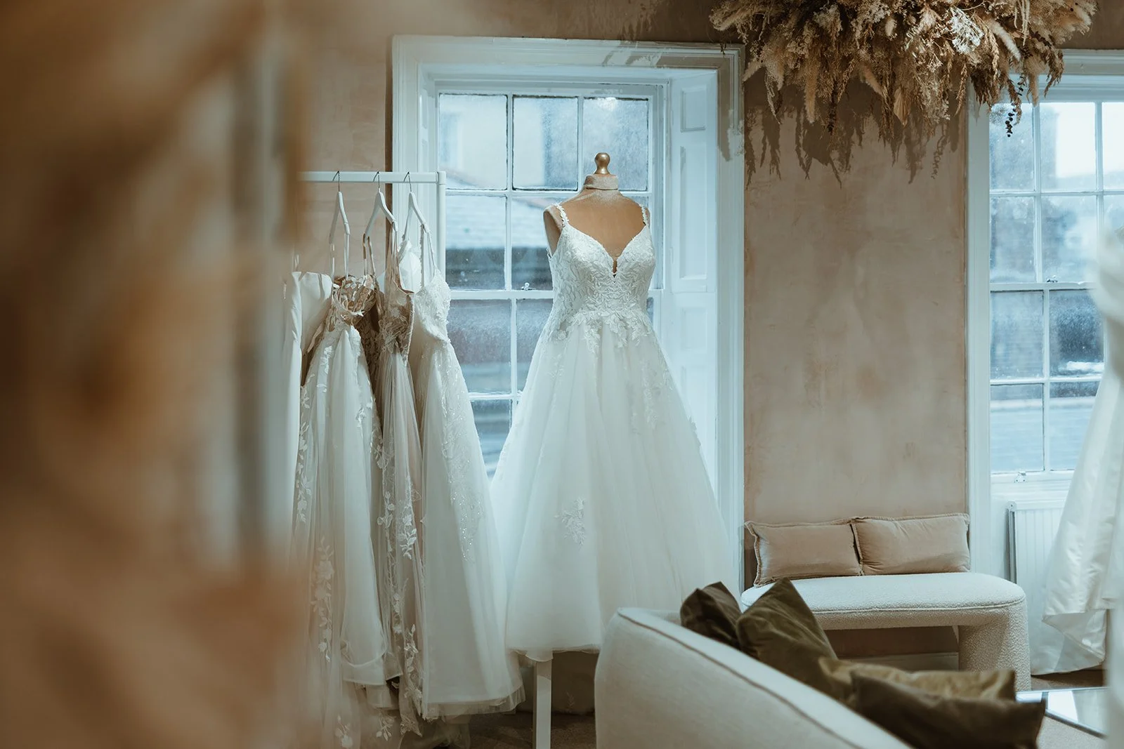 A wedding dress on a mannequin behind a window, with other dresses hanging on a rack nearby, in a cozy bridal boutique.