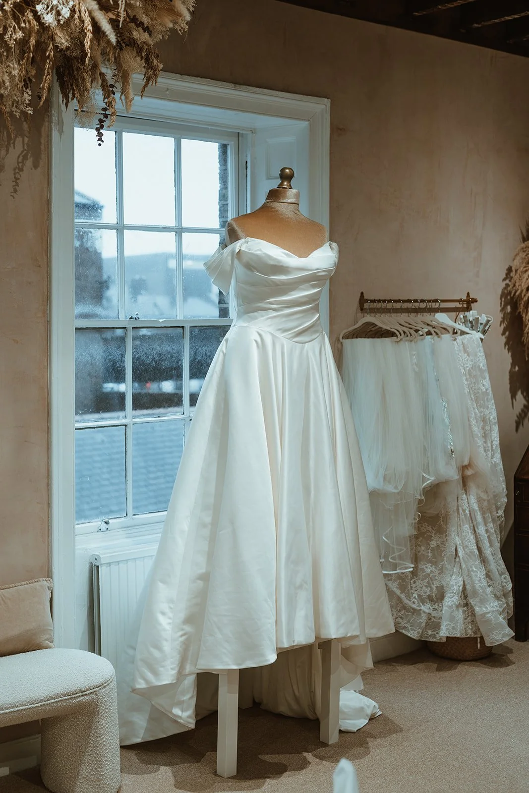 A wedding dress on a mannequin in a room with a window and other wedding dresses hanging on a rack.