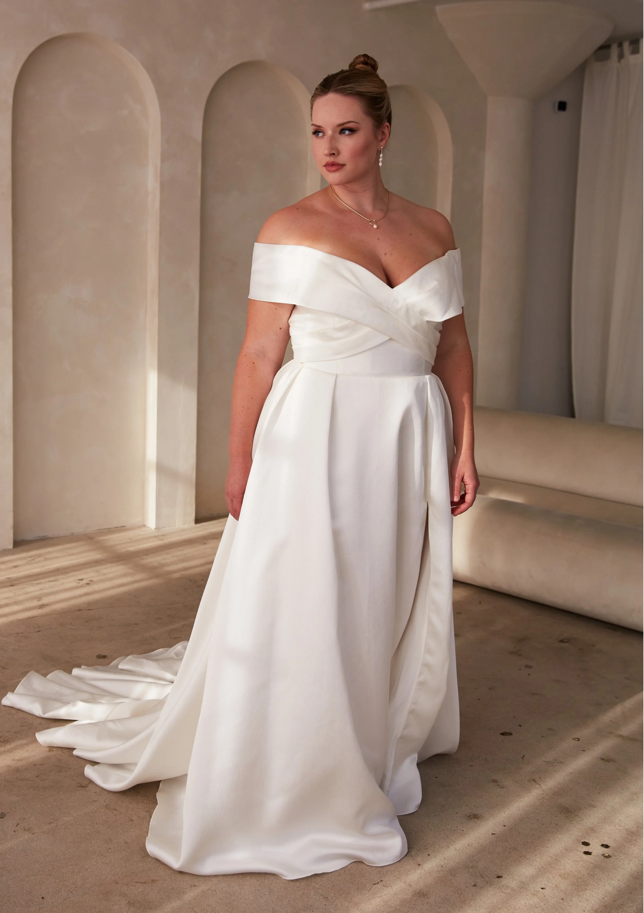 A woman in a white wedding dress standing in a room with light-colored walls and beige curtains.