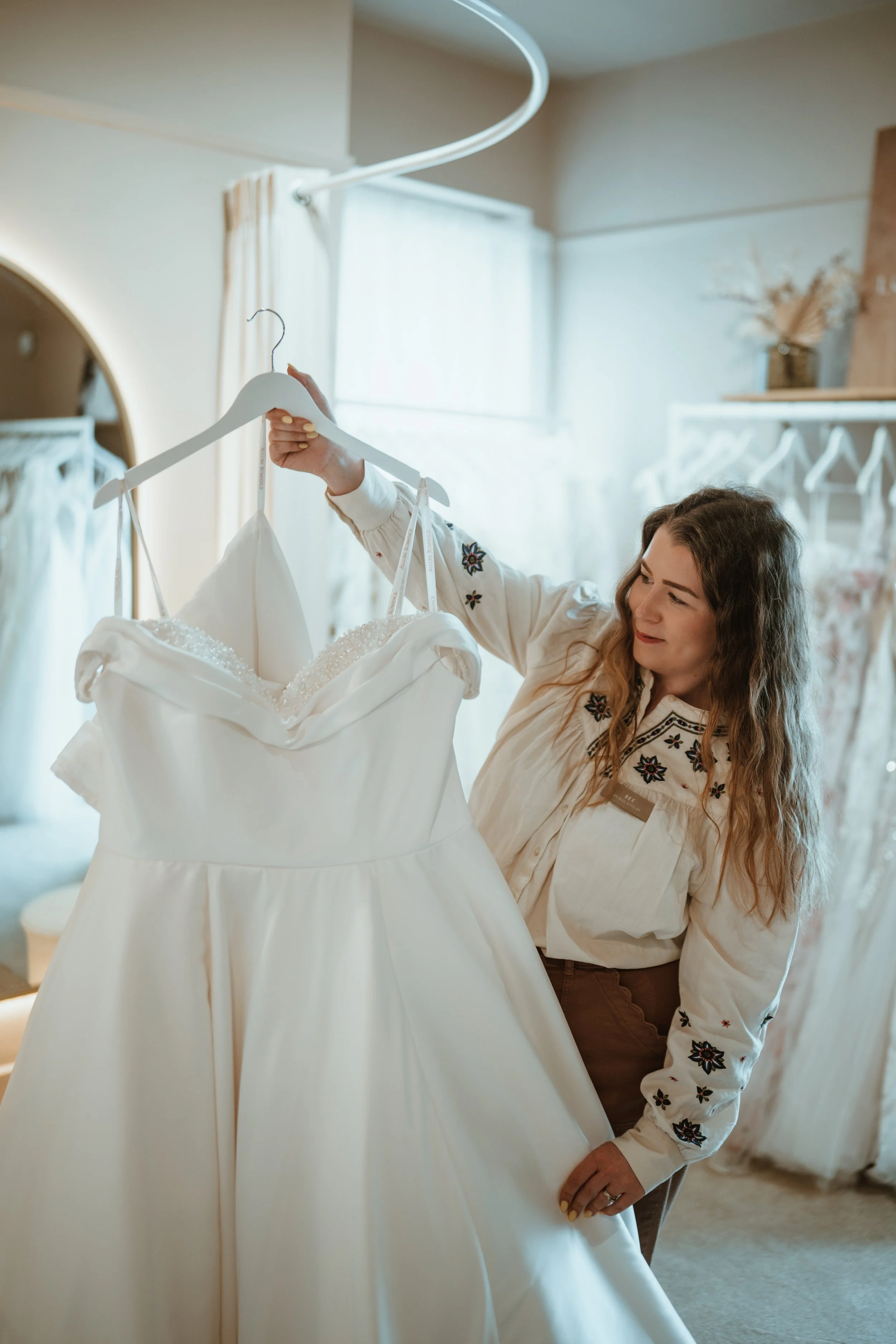 A woman with long curly hair trying on a wedding dress at a bridal boutique, looking at the dress while holding it on a hanger.