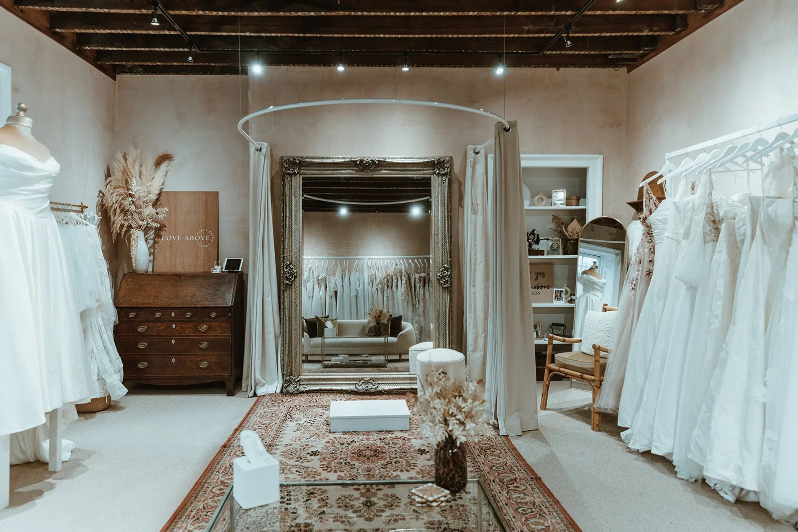 A boutique bridal shop with wedding dresses on racks, a large ornate mirror, a vintage wooden dresser, a cozy seating area, and decorative dried flowers.