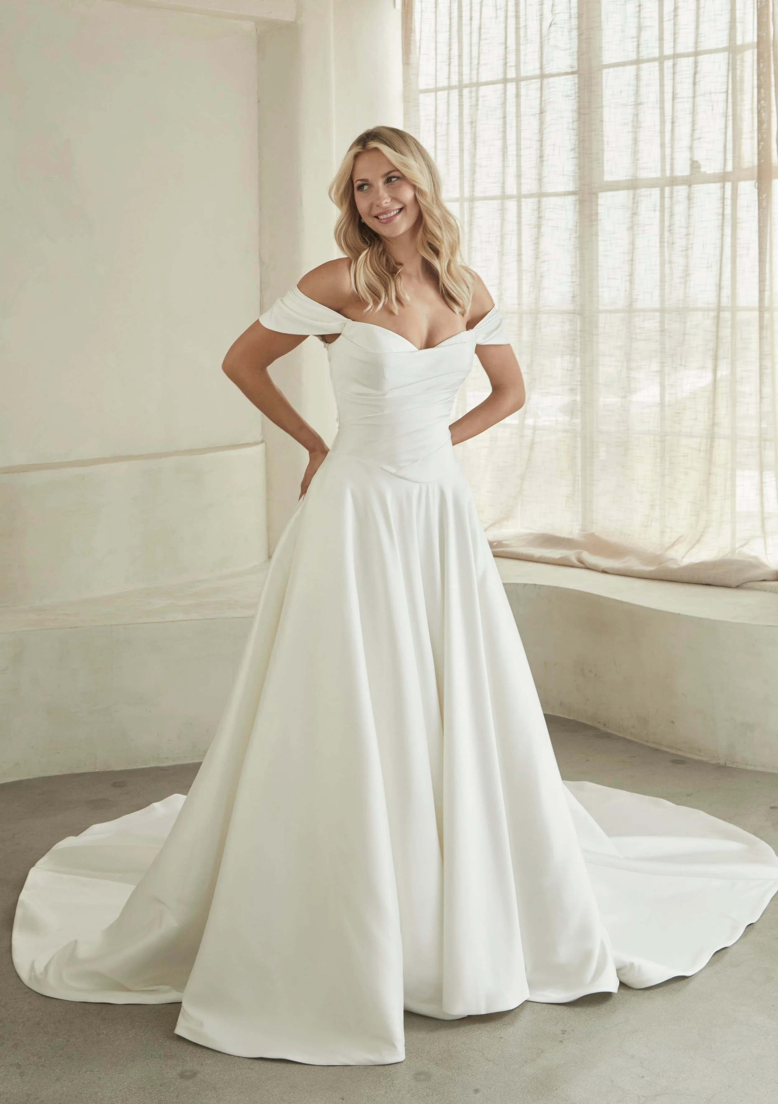 A woman in a white off-shoulder wedding dress posing indoors near a large window with sheer curtains, smiling and looking to her left.