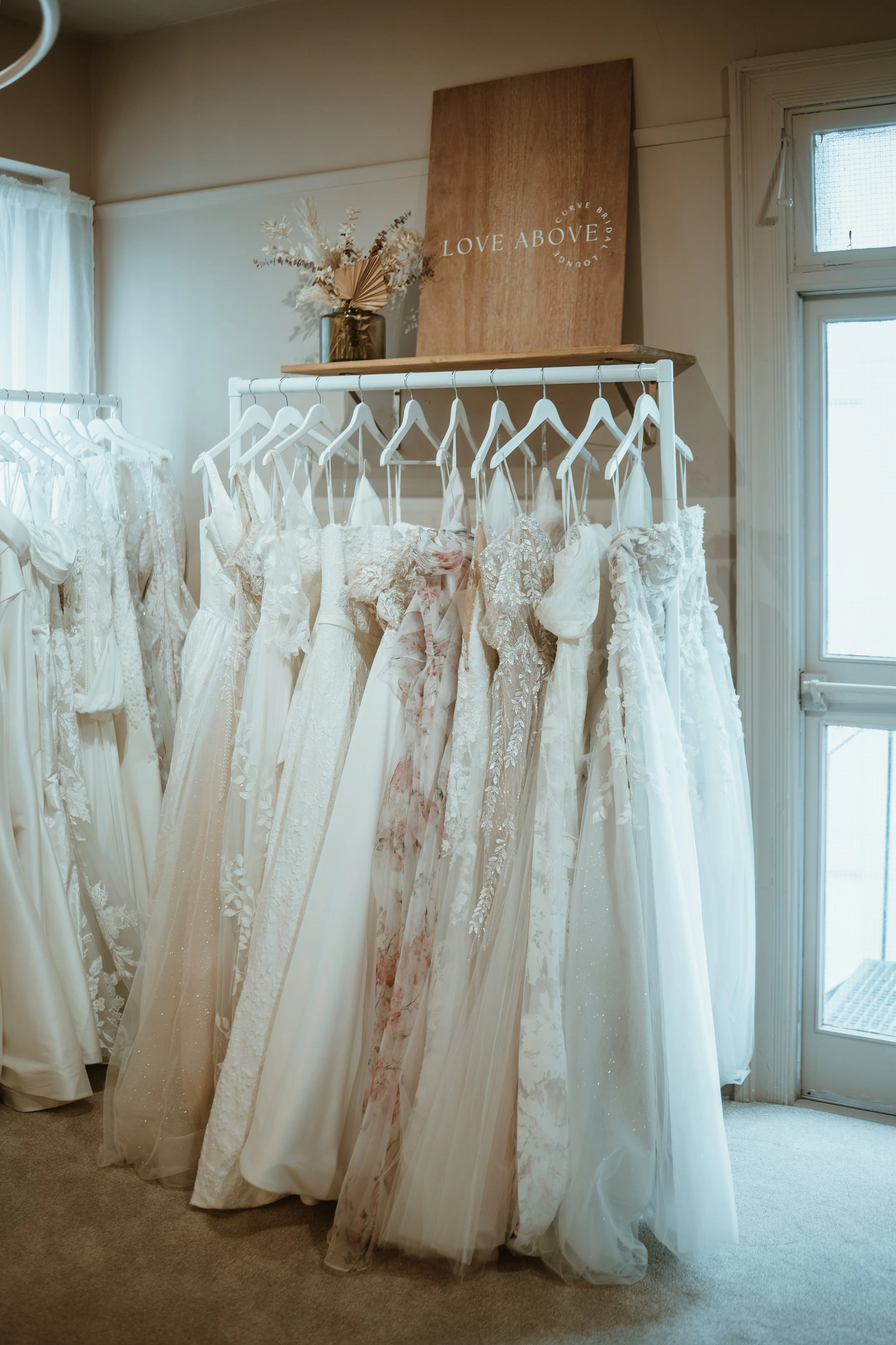 A display of various wedding dresses hanging on a white clothing rack in a boutique, with a wooden sign that reads 'Love Above' above the dresses.