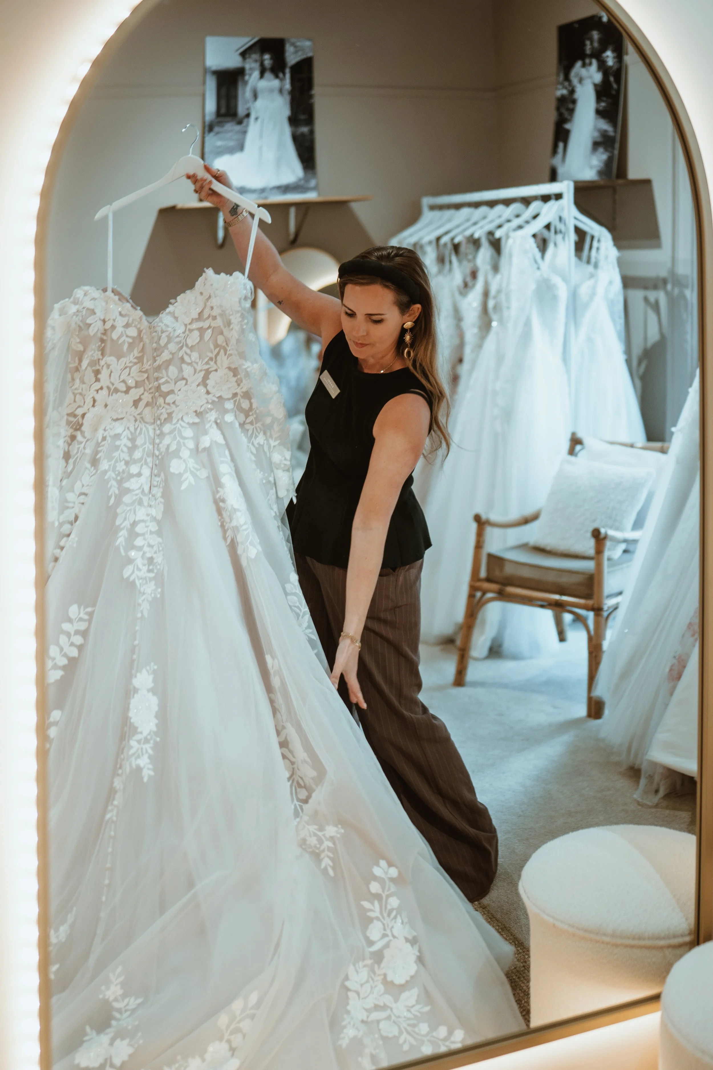 Woman looking at a wedding dress in a dressing room with wedding dresses hanging behind her.
