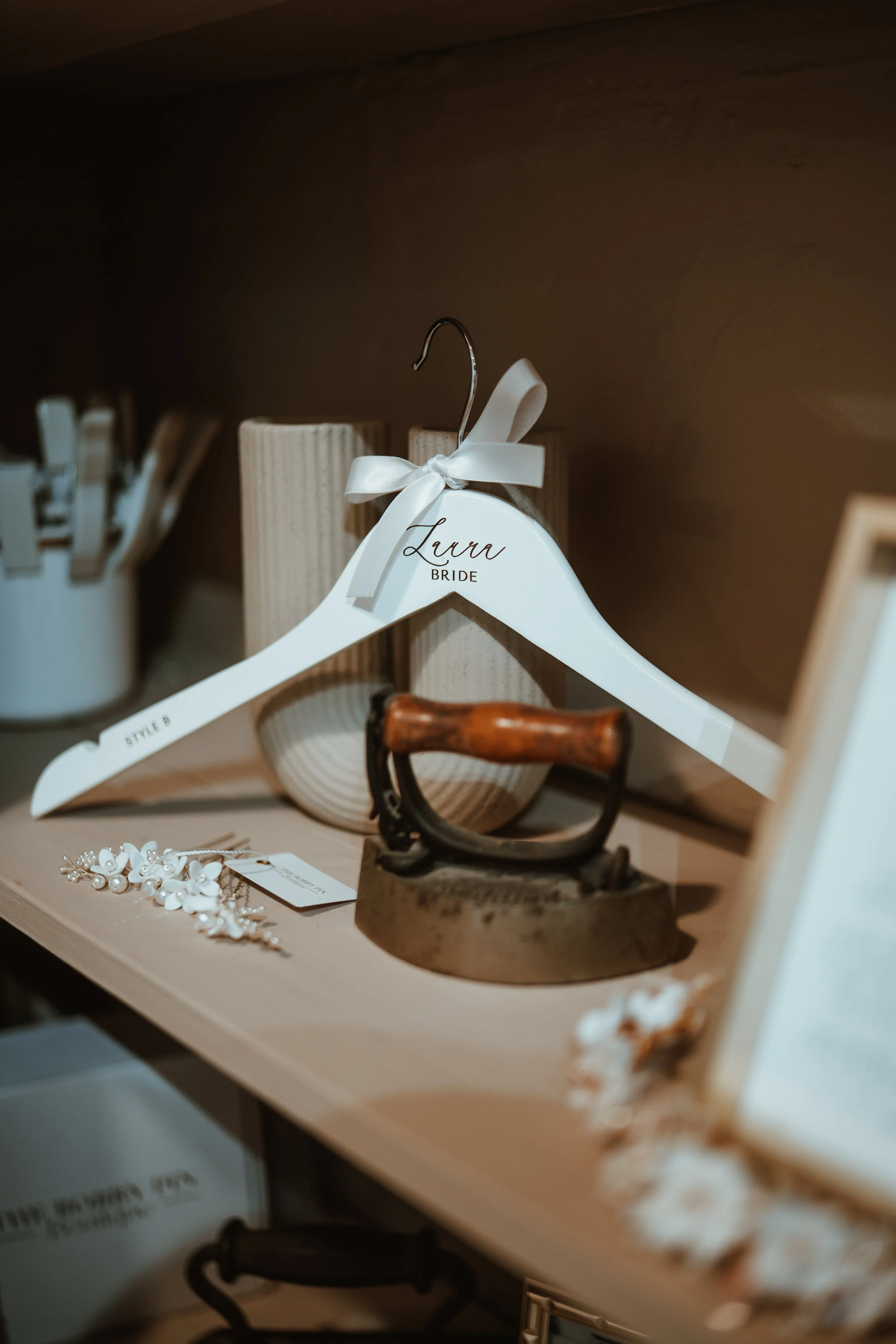A white hanger labeled "Laura BRIDE" hanging on a wooden shelf, with decorative jewelry including pearls and floral accents below it, and various other items on the shelf such as a vintage iron and a candle holder.