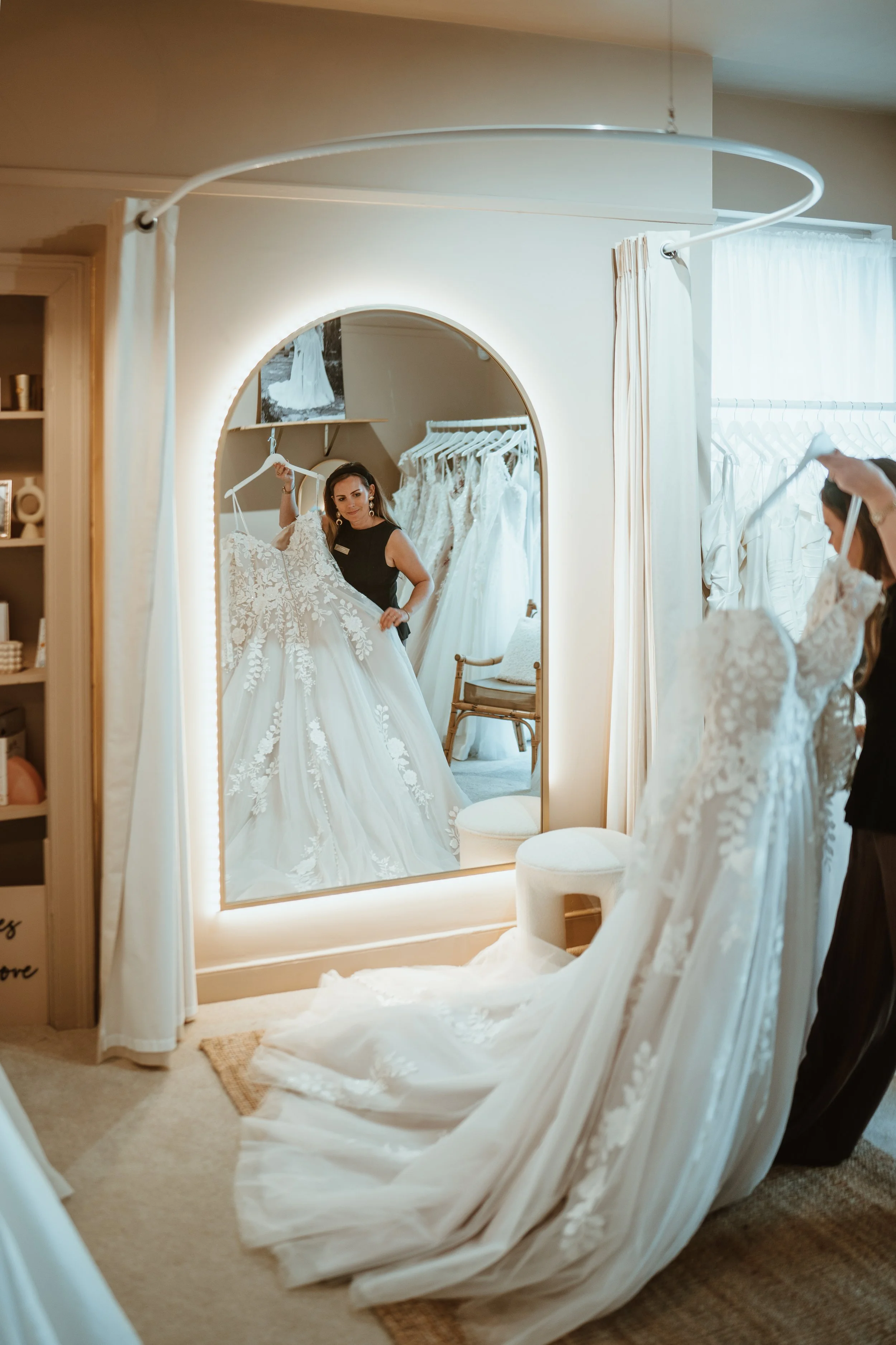 A woman trying on a wedding dress in front of a mirror, with the dress flowing onto the floor and draping over a chair, surrounded by bridal gowns.