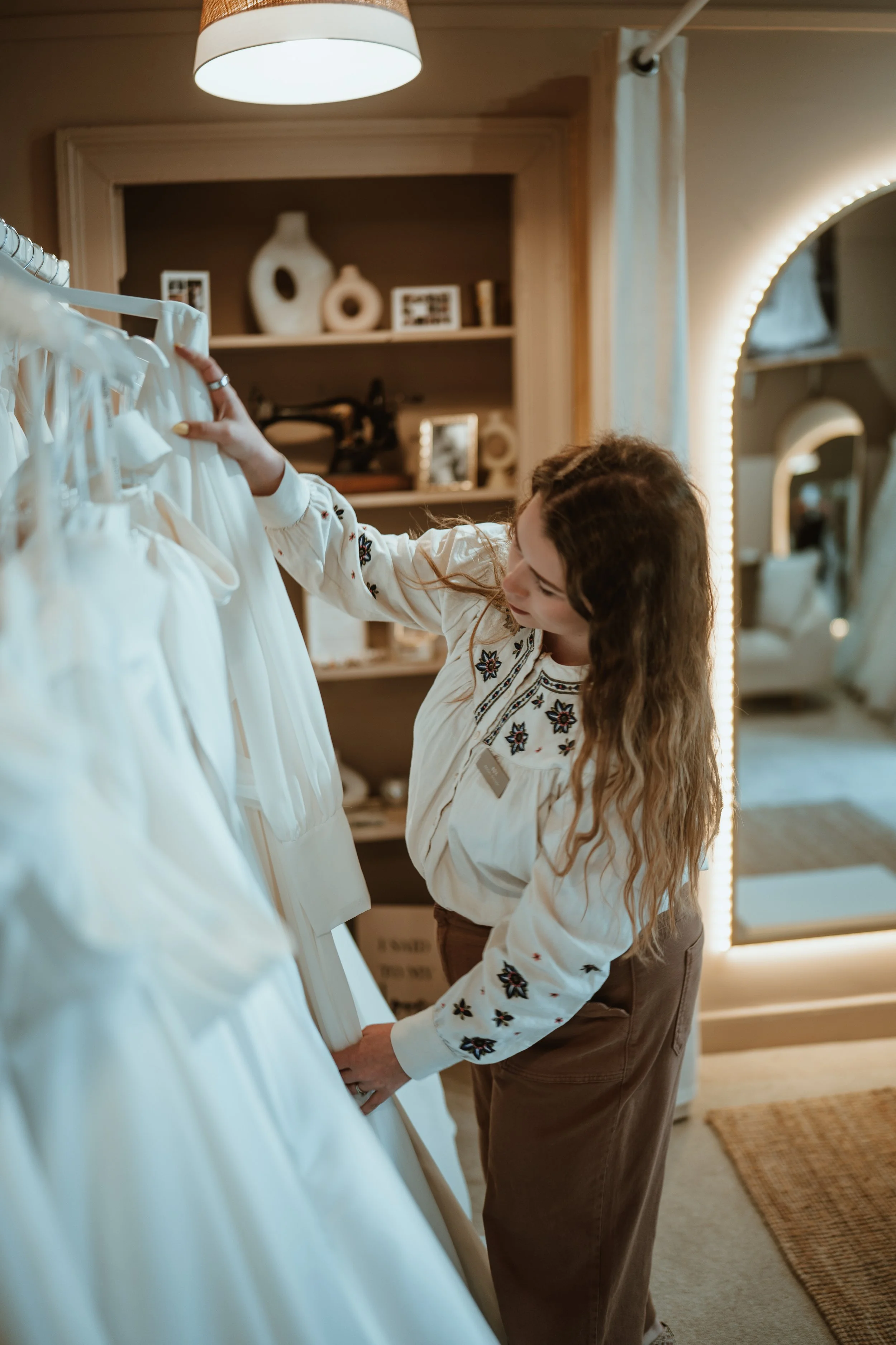 A woman with long wavy hair and a girl with long hair, both wearing embroidered white blouses, are browsing white dresses on a clothing rack in a warmly lit room with a large mirror and decorative shelves in the background.