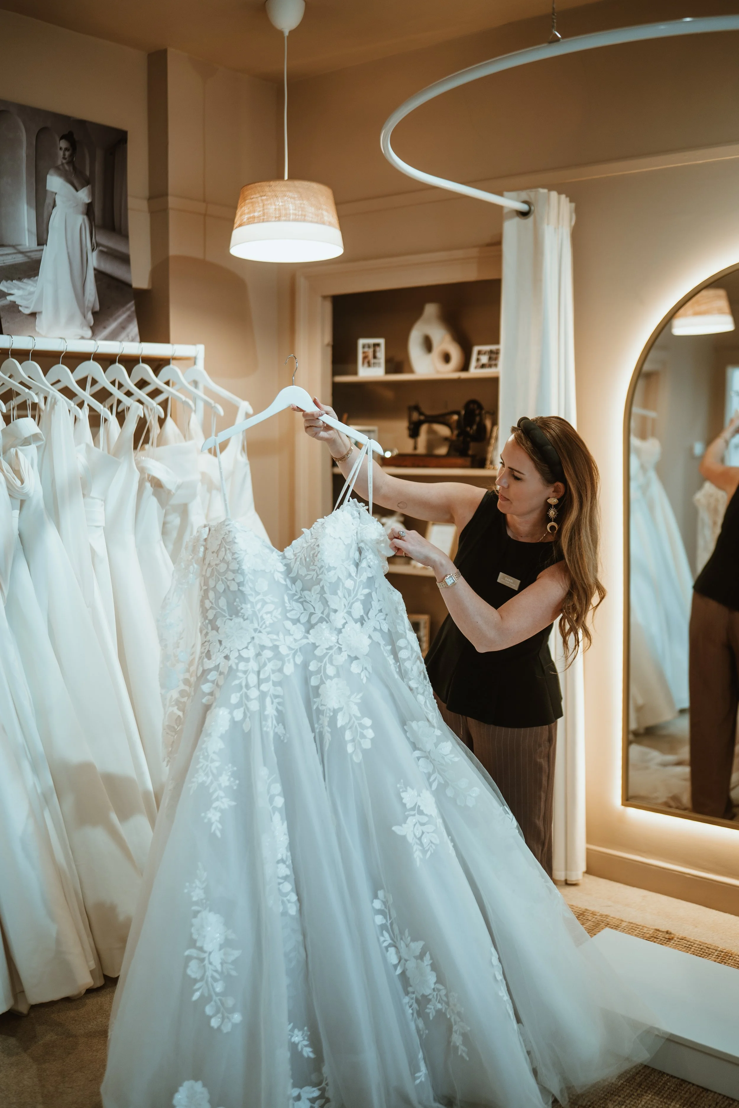 A woman shopping for wedding dresses in a bridal boutique, looking at a white wedding gown with floral embroidery while holding it on a hanger.