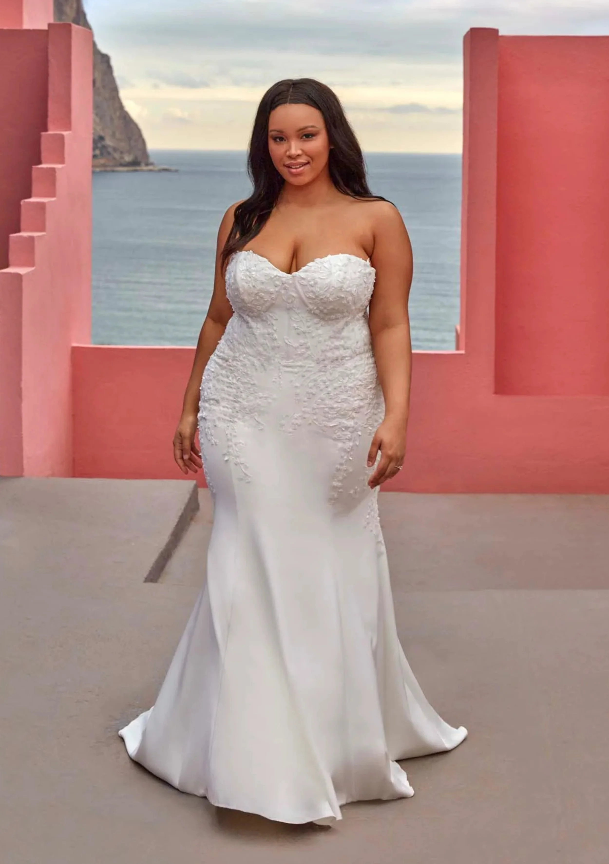 A woman in a white wedding dress standing on a balcony with pink walls, overlooking the ocean and a rocky cliff in the background.