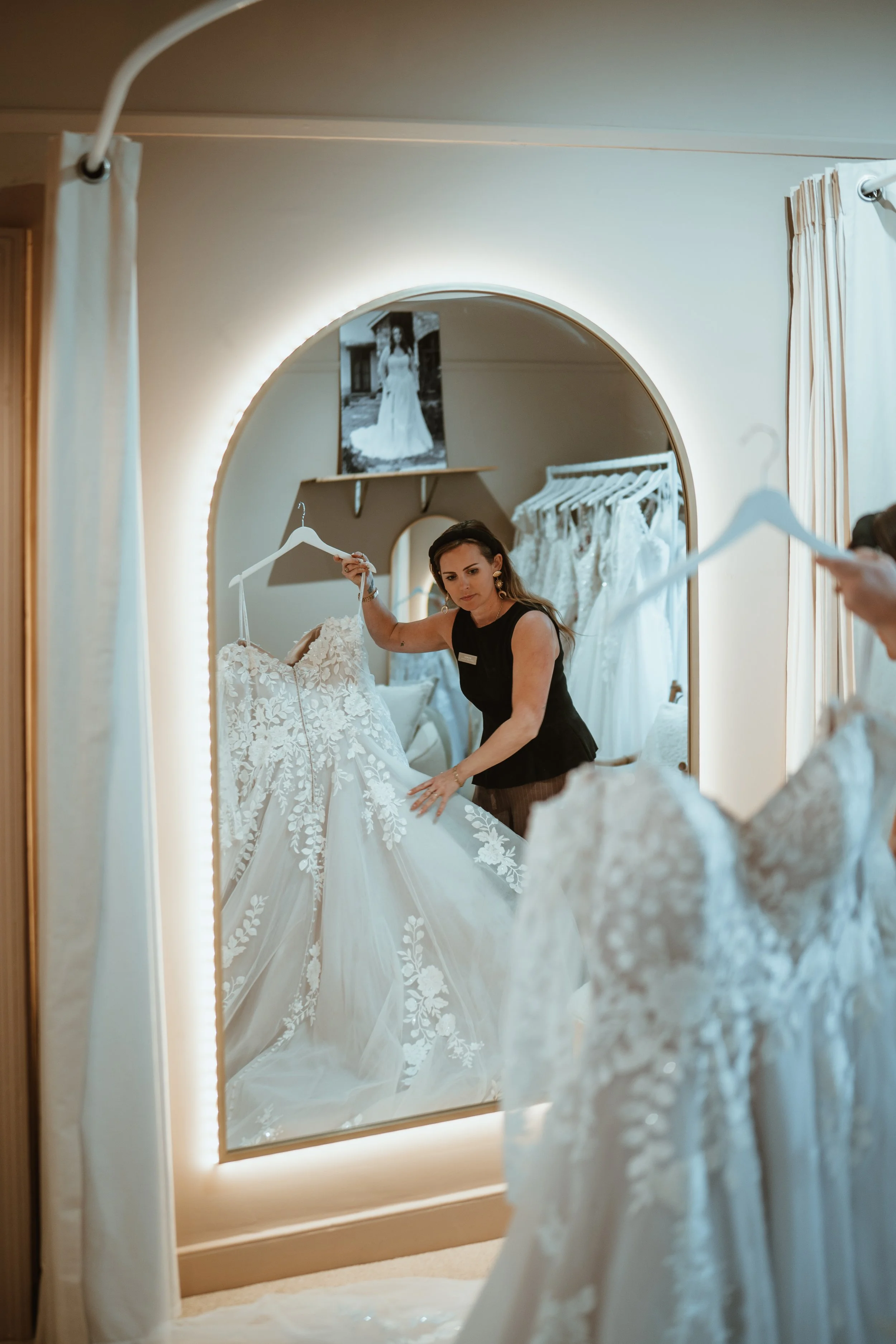 Woman shopping for a wedding dress, looking at a white lace wedding gown on a hanger in a fitting room, reflected in a large mirror.