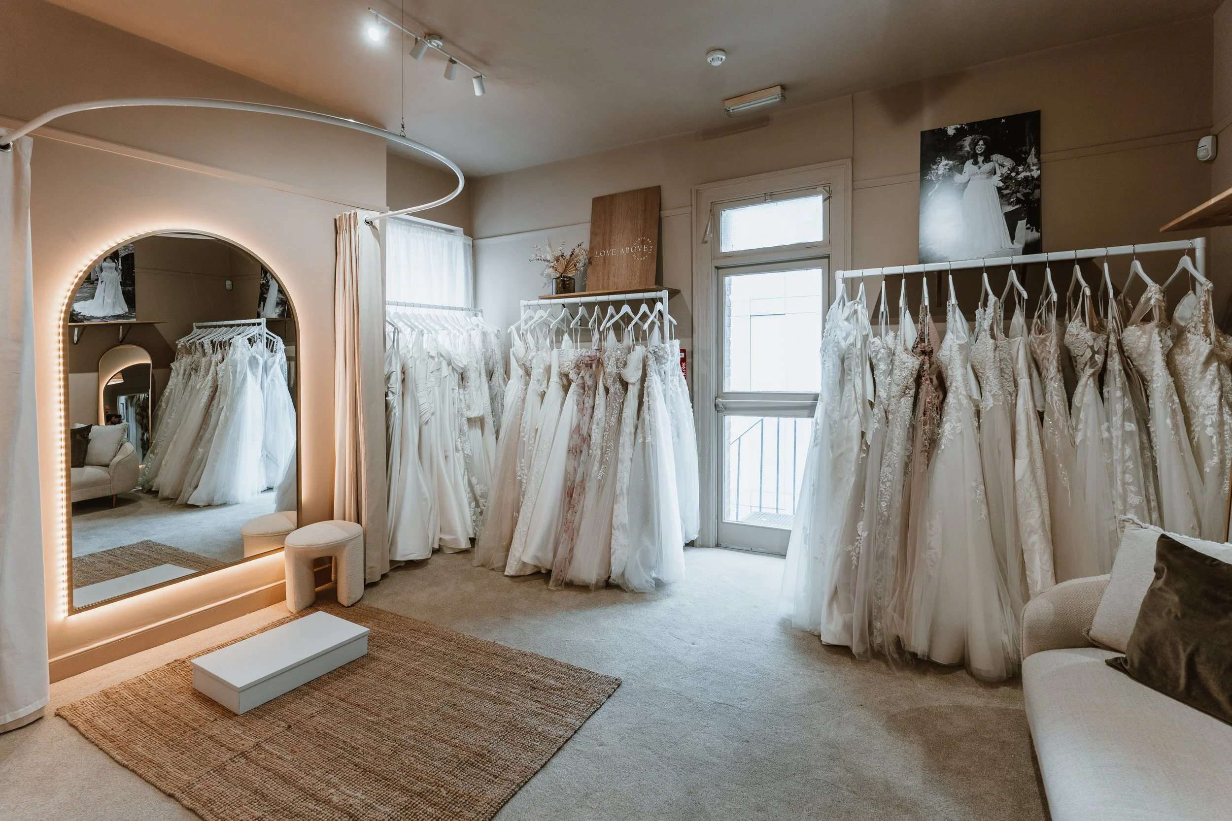 A bridal shop fitting room with wedding dresses hanging on racks, a large illuminated mirror, and a cozy seating area near windows.