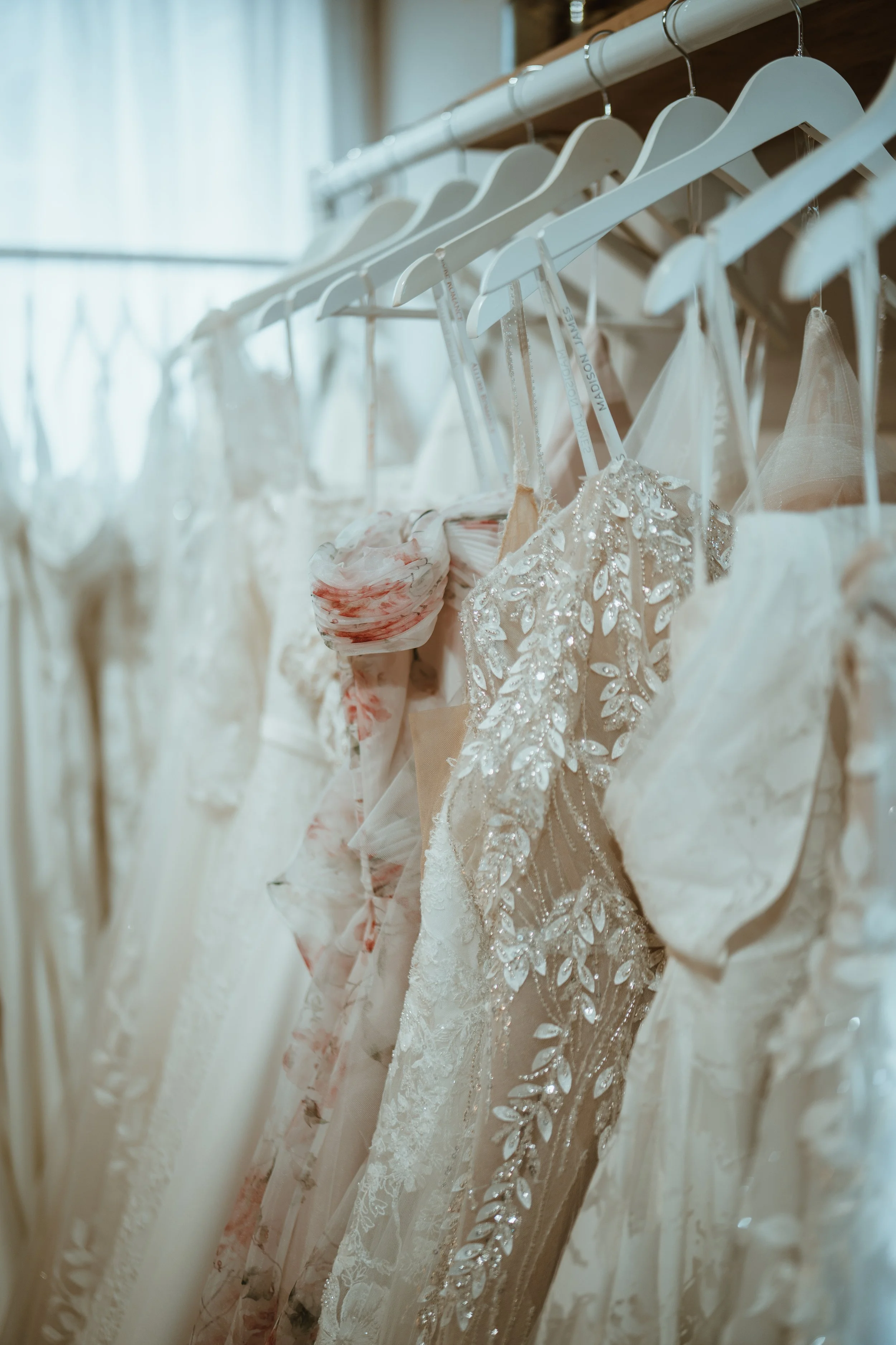 A row of wedding dresses hanging on white hangers on a metal clothing rack, with a window in the background casting natural light.