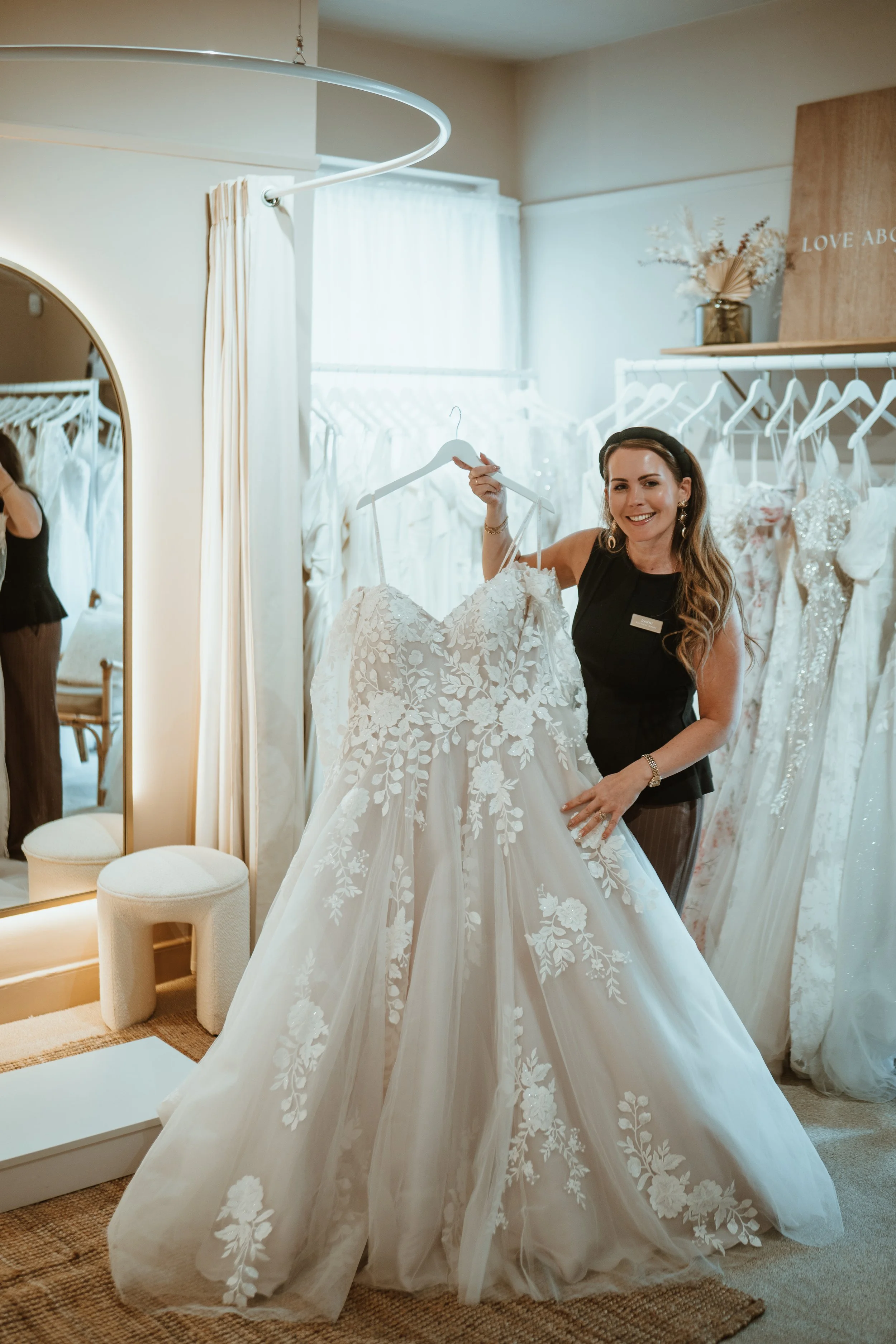 A woman is inside a bridal boutique holding up a white wedding dress with floral lace details, surrounded by similar wedding gowns hanging on a clothing rack.
