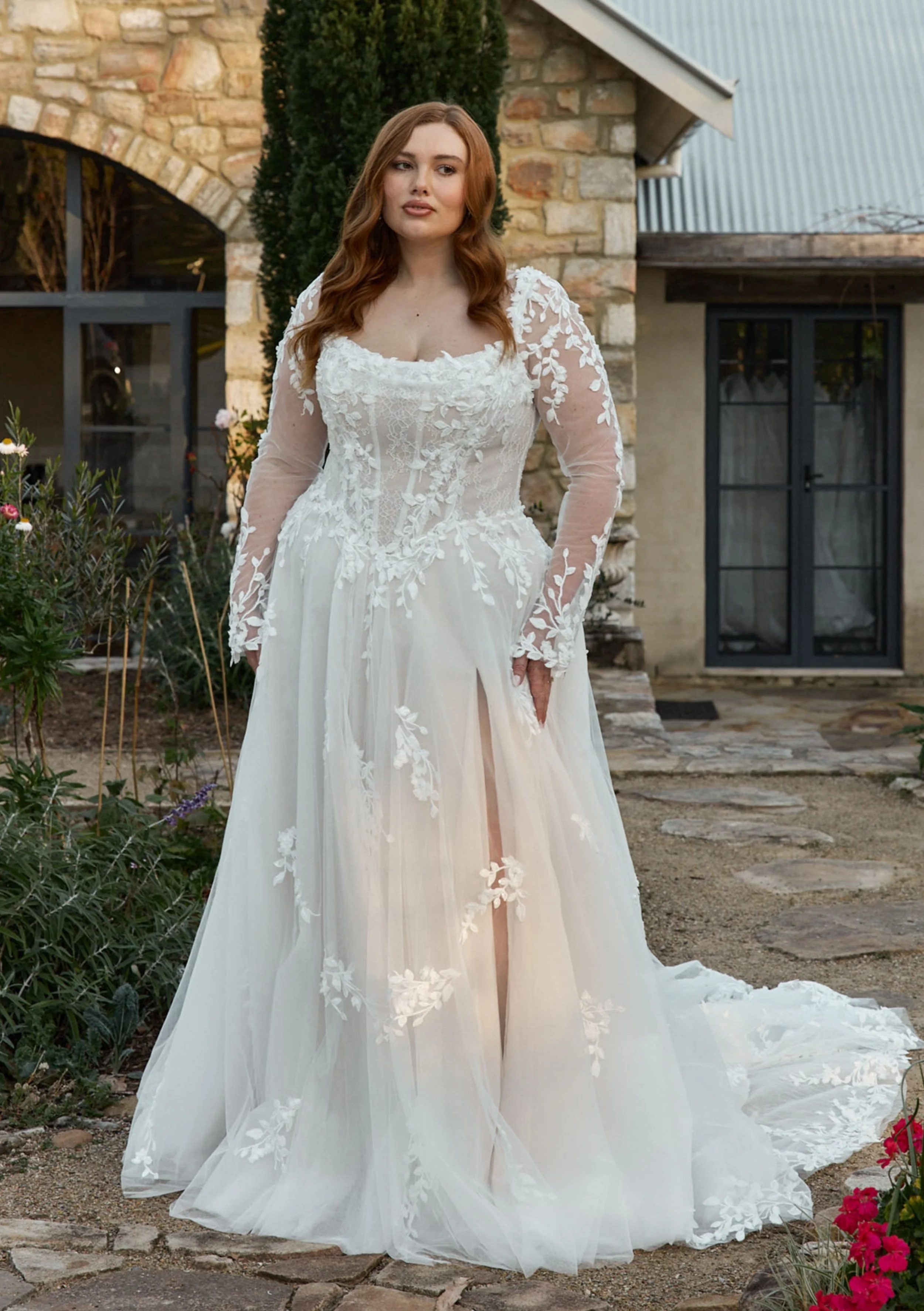 A woman in a white wedding dress with lace details standing outdoors next to a stone building with a garden and flowers.