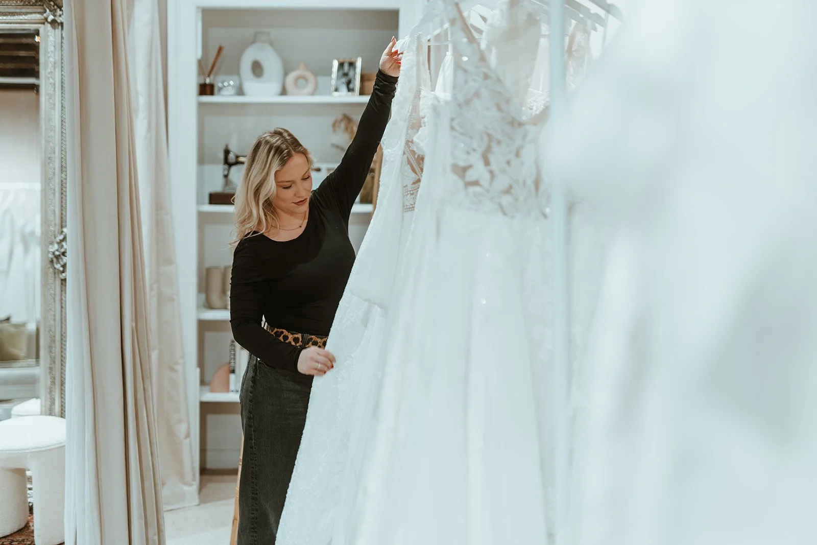 A woman with blonde hair is shopping for wedding dresses in a boutique, browsing through white wedding gowns hanging on a rack.