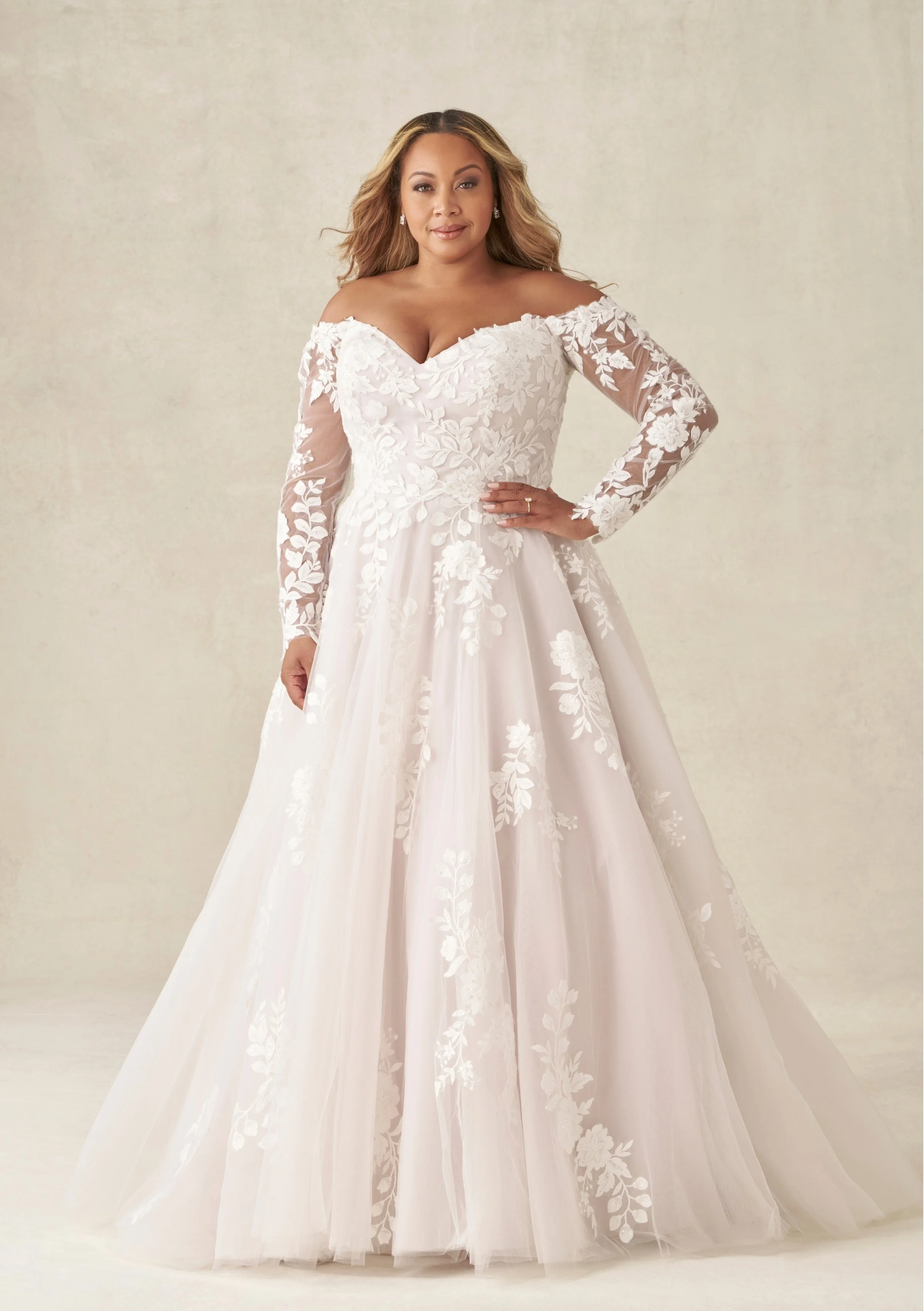 A woman in a white wedding dress with off-the-shoulder lace sleeves and floral embroidery, standing against a neutral background.