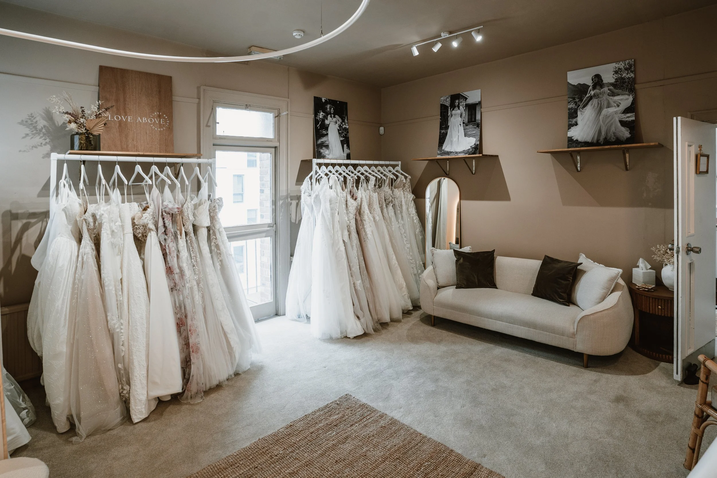 A bridal boutique with a collection of white wedding dresses hanging on a rack by a window. Black and white photographs of women in wedding gowns adorn the walls. There is a beige sofa with dark cushions, a mirror, and decorative shelves with flowers and art.