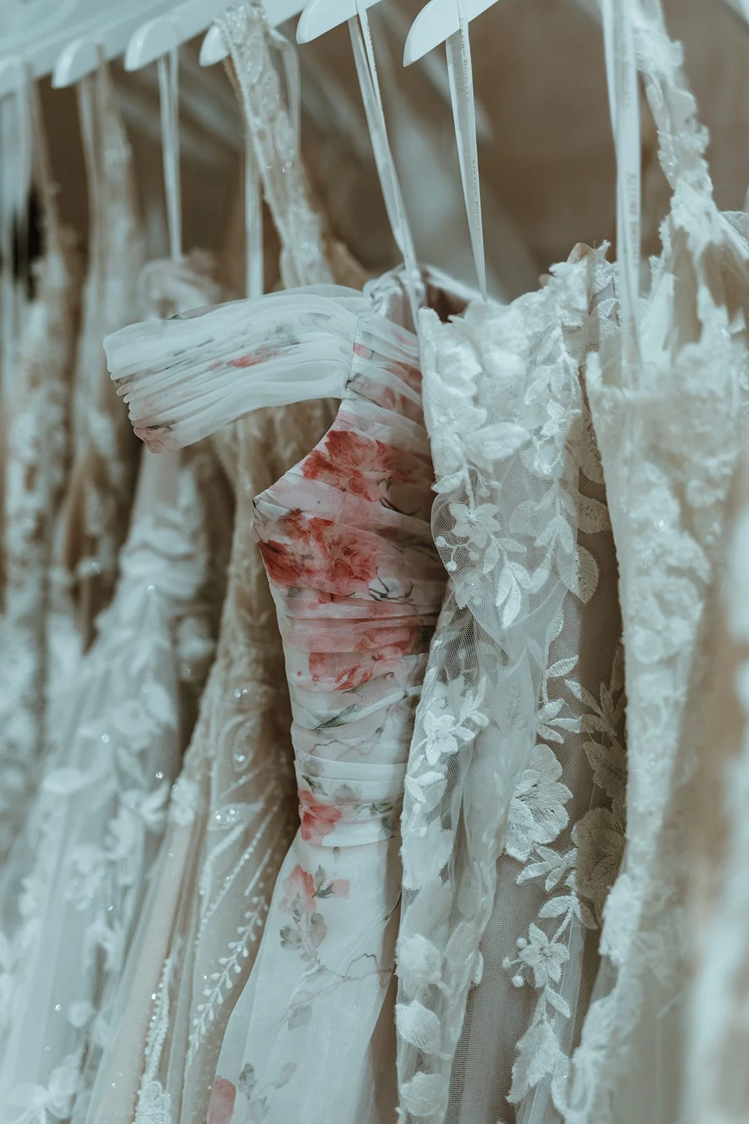 A row of white and cream wedding dresses with lace and floral details hanging on white hangers.