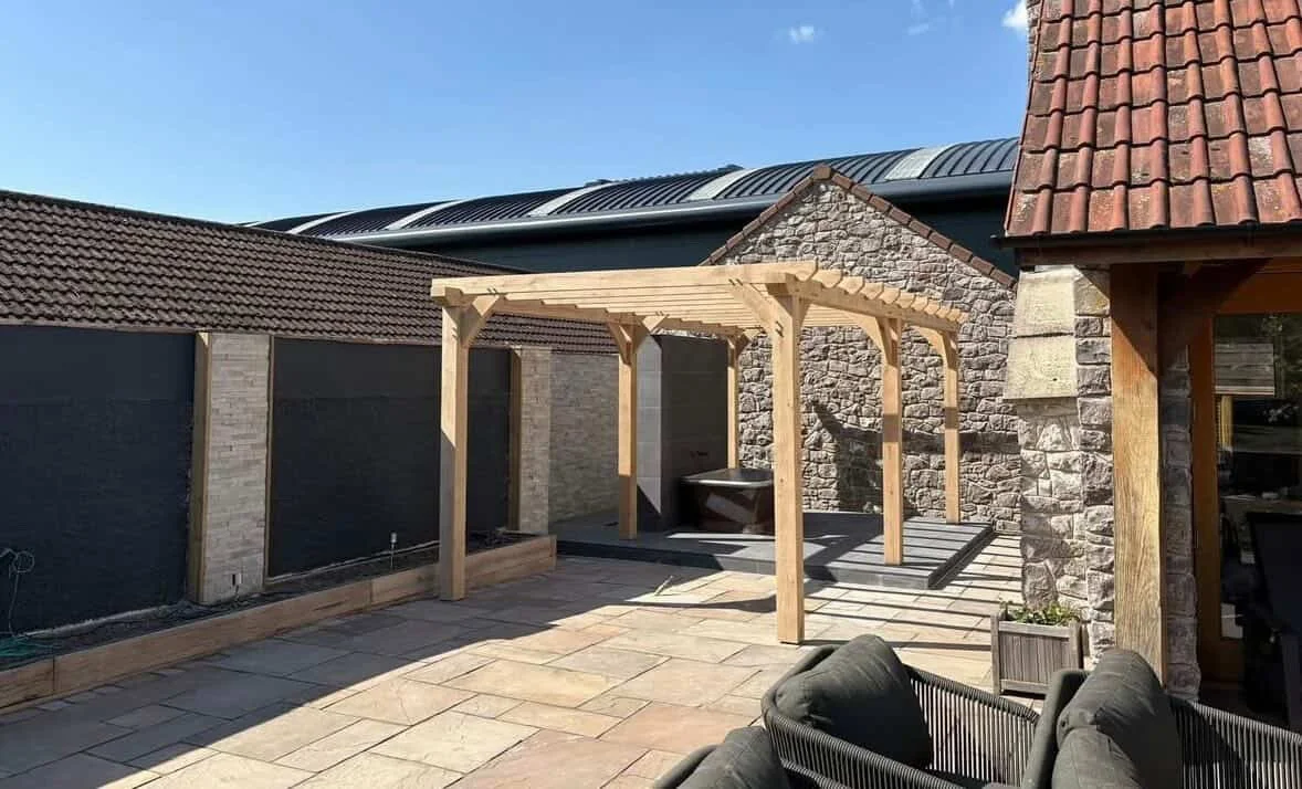 Backyard patio area with a wooden pergola frame, stone and brick walls, tiled patio floor, and outdoor seating.