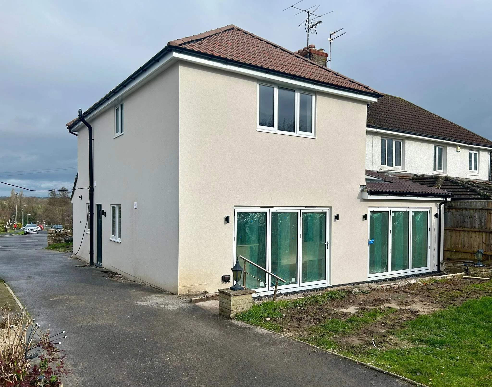 A newly renovated two-story house with white exterior walls, brown tiled roof, and large glass sliding doors on the ground floor. The property is situated in a suburban neighborhood with a partially paved driveway and some patches of grass and soil i