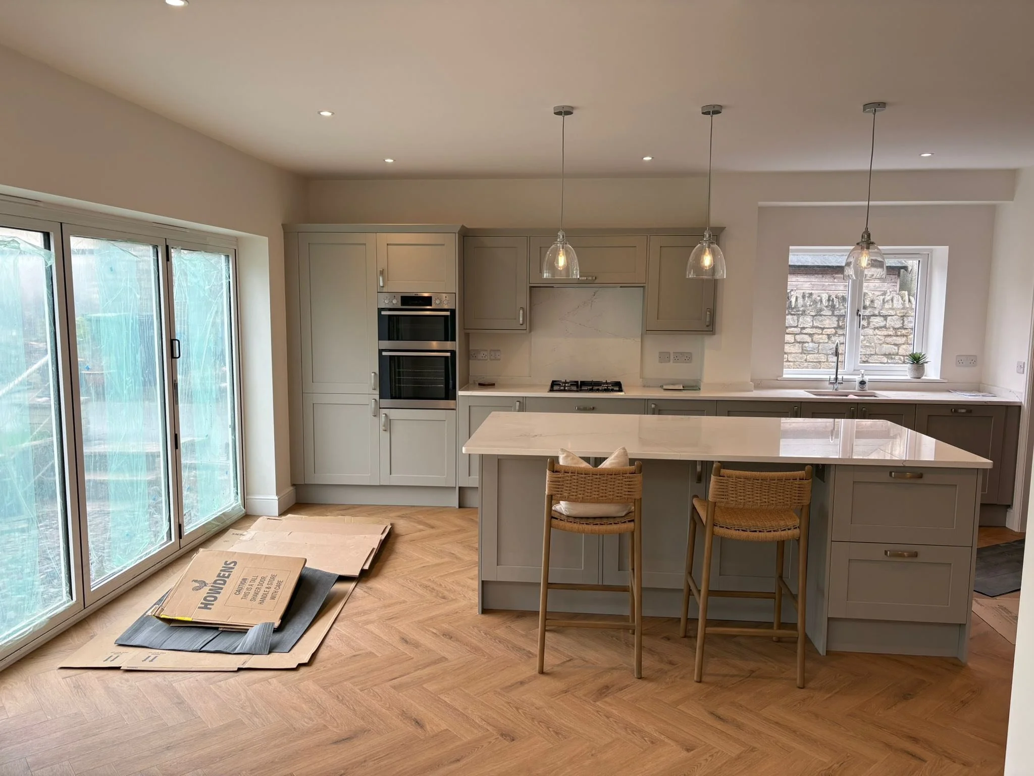 Modern kitchen with an island, beige cabinets, marble backsplash, and pendant lights, with a sliding glass door and a window overlooking a stone wall.