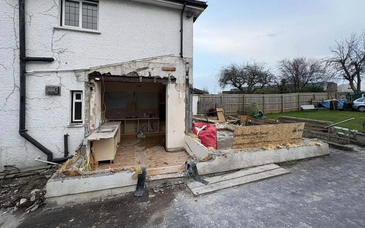 Construction or renovation site of a house with the front section partially demolished, exposing the interior workspace, construction materials, and tools.
