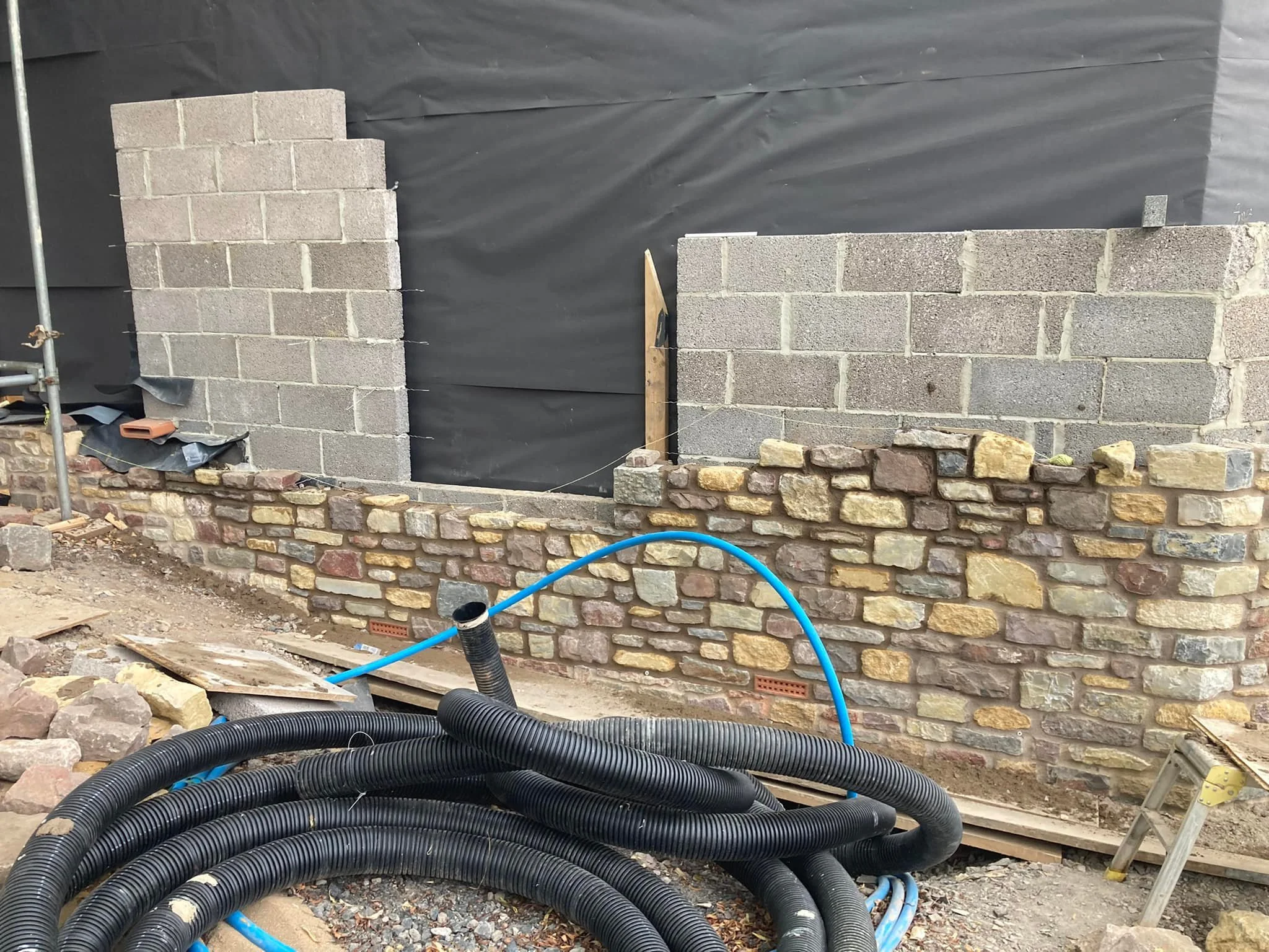 Construction site with brick wall being built, black hoses and blue pipes in foreground, some wall sections made of concrete blocks, scaffolding on the left, black plastic sheeting covering the background.