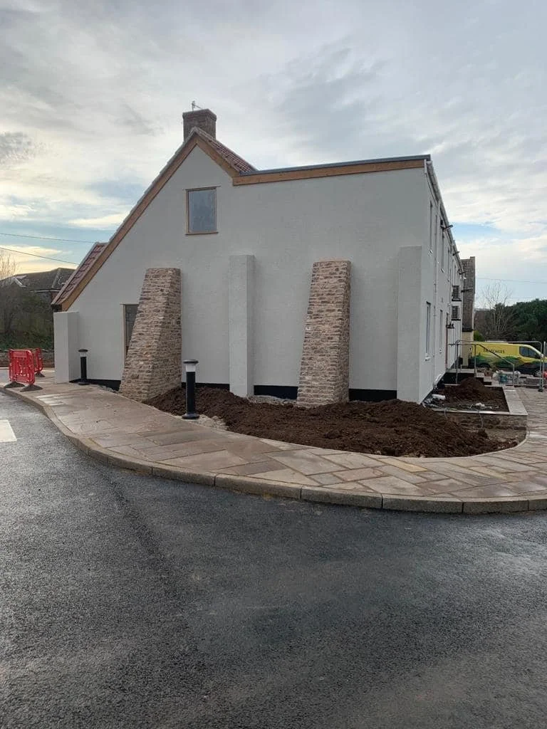 Newly constructed house with white exterior walls, brick accents, and a steep roof, situated on a corner with a paved sidewalk and street, with construction materials and equipment nearby.