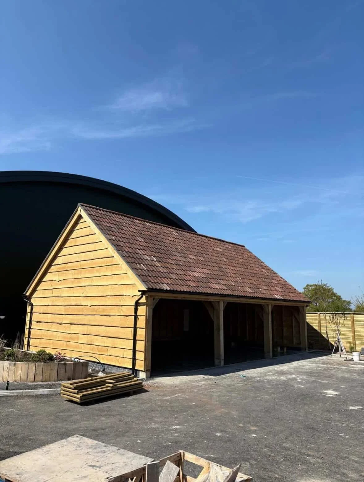 A small wooden garage with a tiled gable roof under construction, surrounded by construction materials and tools, with a blue sky overhead.