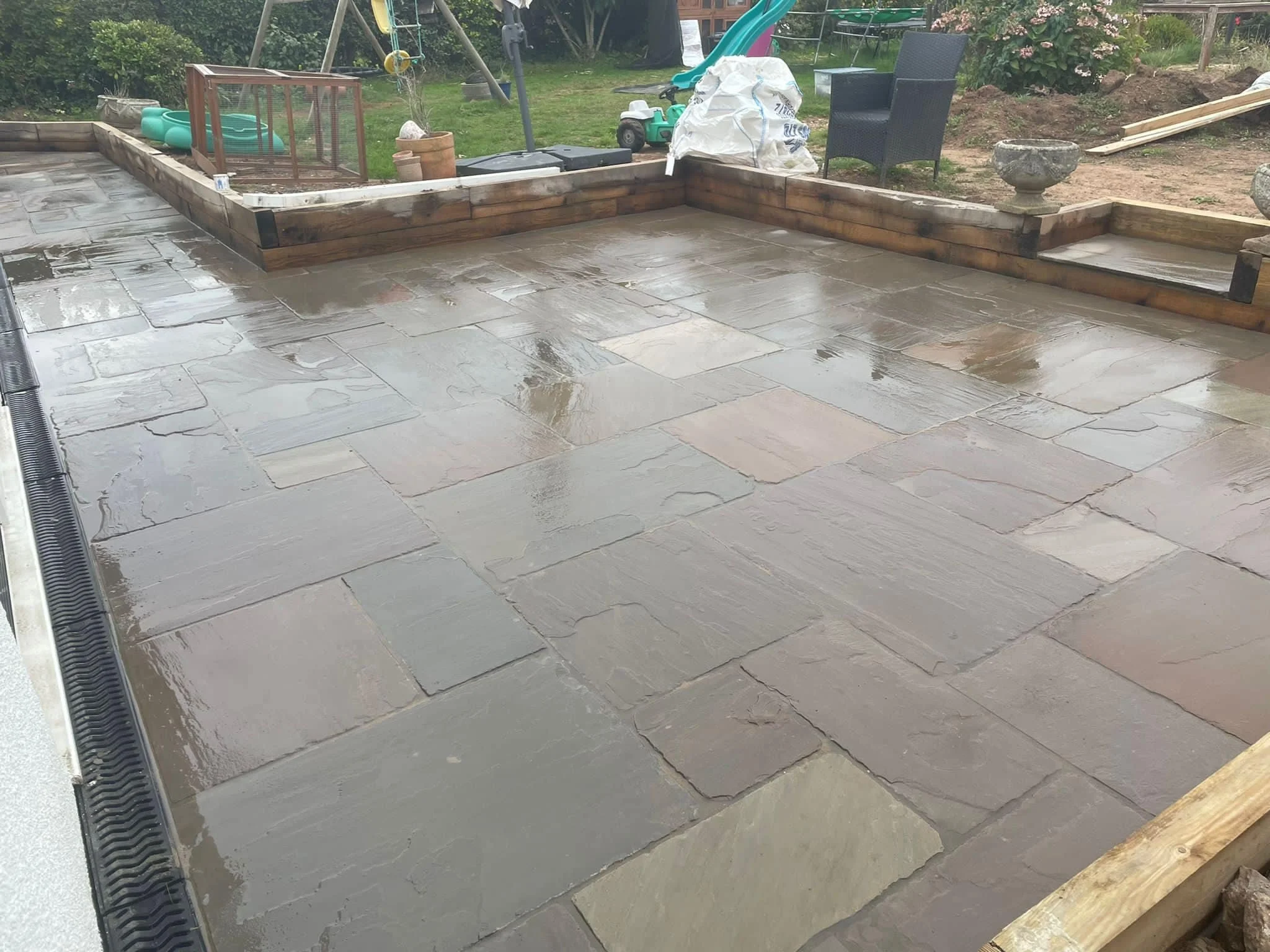 Wet outdoor patio with stone tiles and wooden border, in a backyard with garden tools, plants, and outdoor furniture.