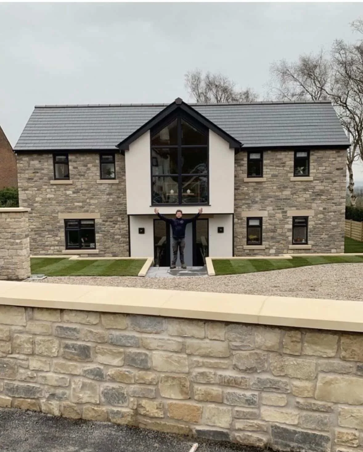 A man standing in front of a large modern stone house with his arms raised up.