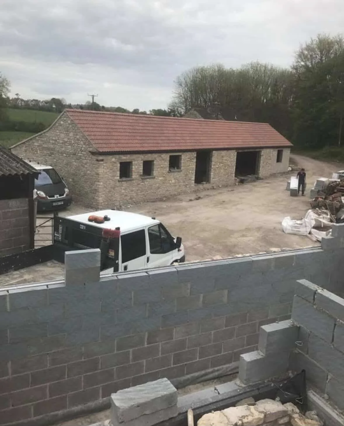 View of a construction site with a stone building, a white van, and a partially built brick wall in the foreground, with trees and a cloudy sky in the background.