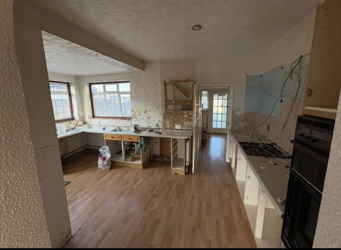 Kitchen undergoing renovation with exposed counter cabinets, missing appliances, damaged walls, and construction wires hanging.