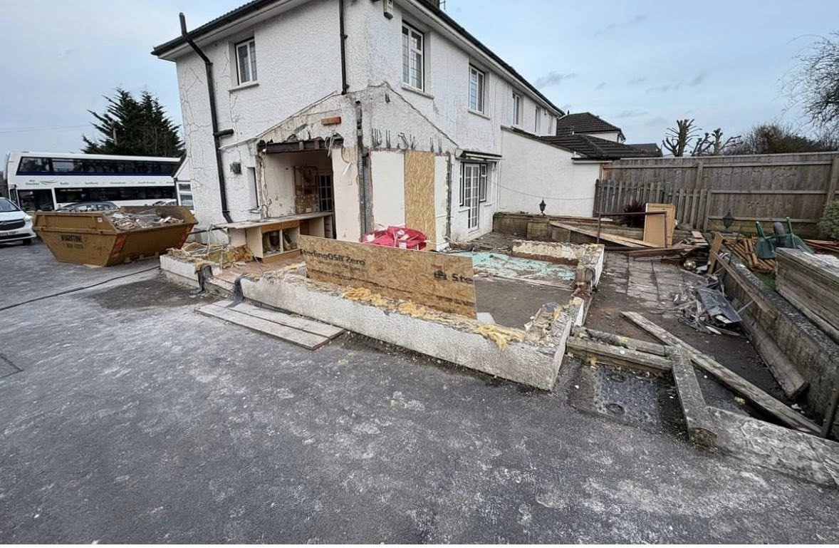 A house under construction or renovation with part of the exterior wall removed, construction debris, wooden planks, and a skip container filled with debris in front.
